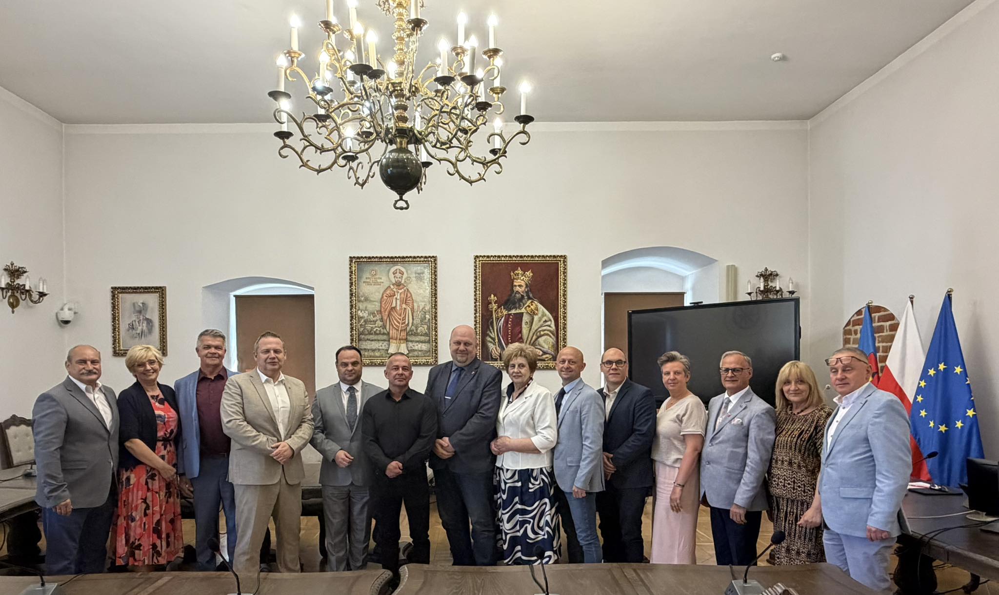 Grupa kobiet i mężczyzn stoi w reprezentacyjnej sali z żyrandolem. W tle widoczne obrazy, ekran oraz flagi Polski i Unii Europejskiej. Uczestnicy pozują do wspólnego zdjęcia podczas oficjalnego spotkania.