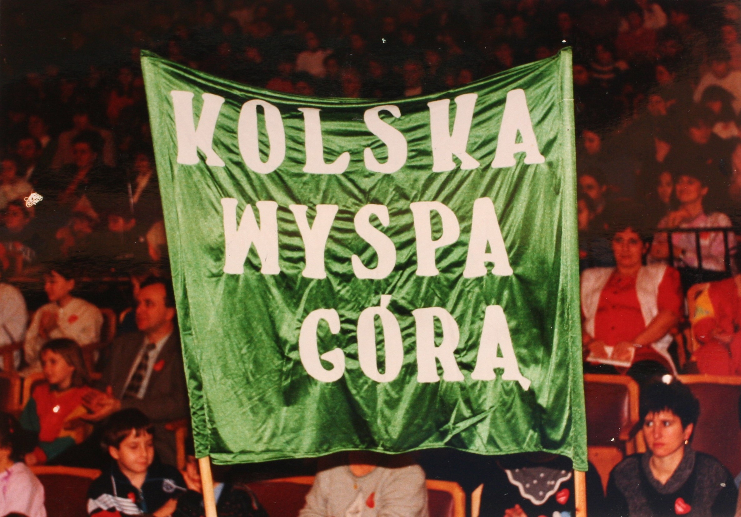 Zdjęcie przedstawia zielony baner z napisem KOLSKA WYSPA GÓRA” trzymany w tłumie ludzi siedzących na widowni. W tle siedzące osoby na widowni.