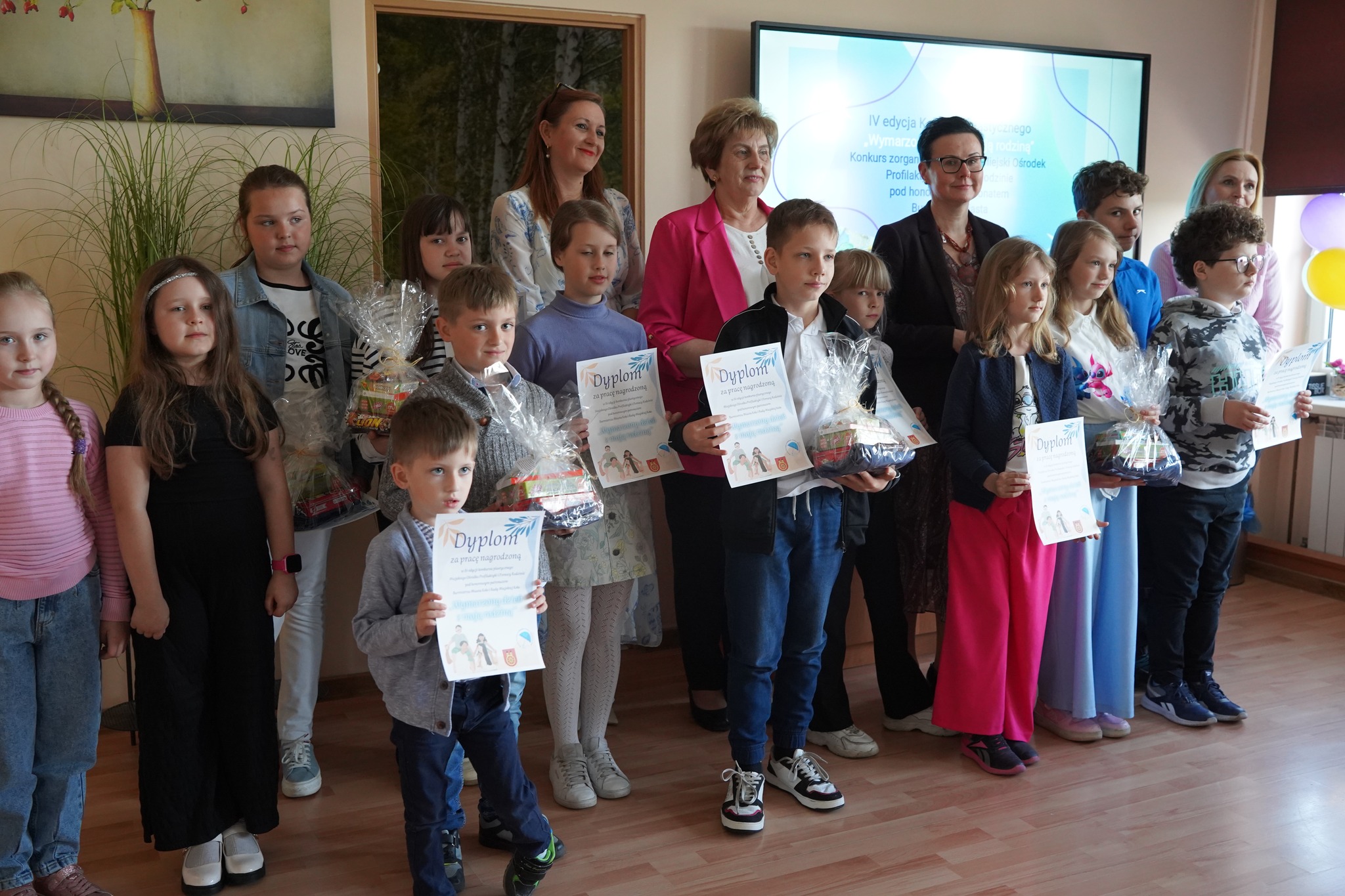 Zdjęcie przedstawia grupę dzieci i dorosłych podczas rozdania nagród – dzieci trzymają dyplomy i upominki, a w tle widać ekran z informacją o konkursie rodzinnym.