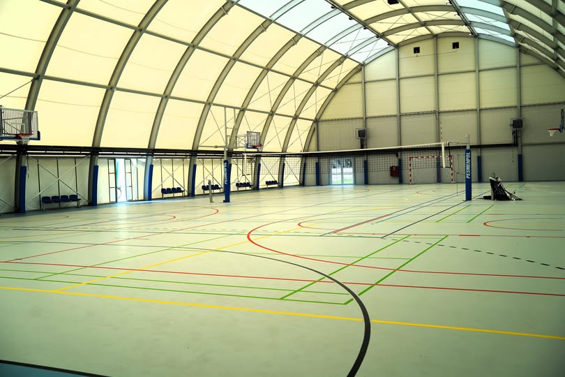 Wnętrze pustej hali sportowej z łukowym, jasnym dachem namiotowym na stalowych łukach. Szara podłoga z kolorowymi liniami boisk, rozstawiona siatka do siatkówki, kosze do koszykówki, przy ścianie rząd krzesełek.