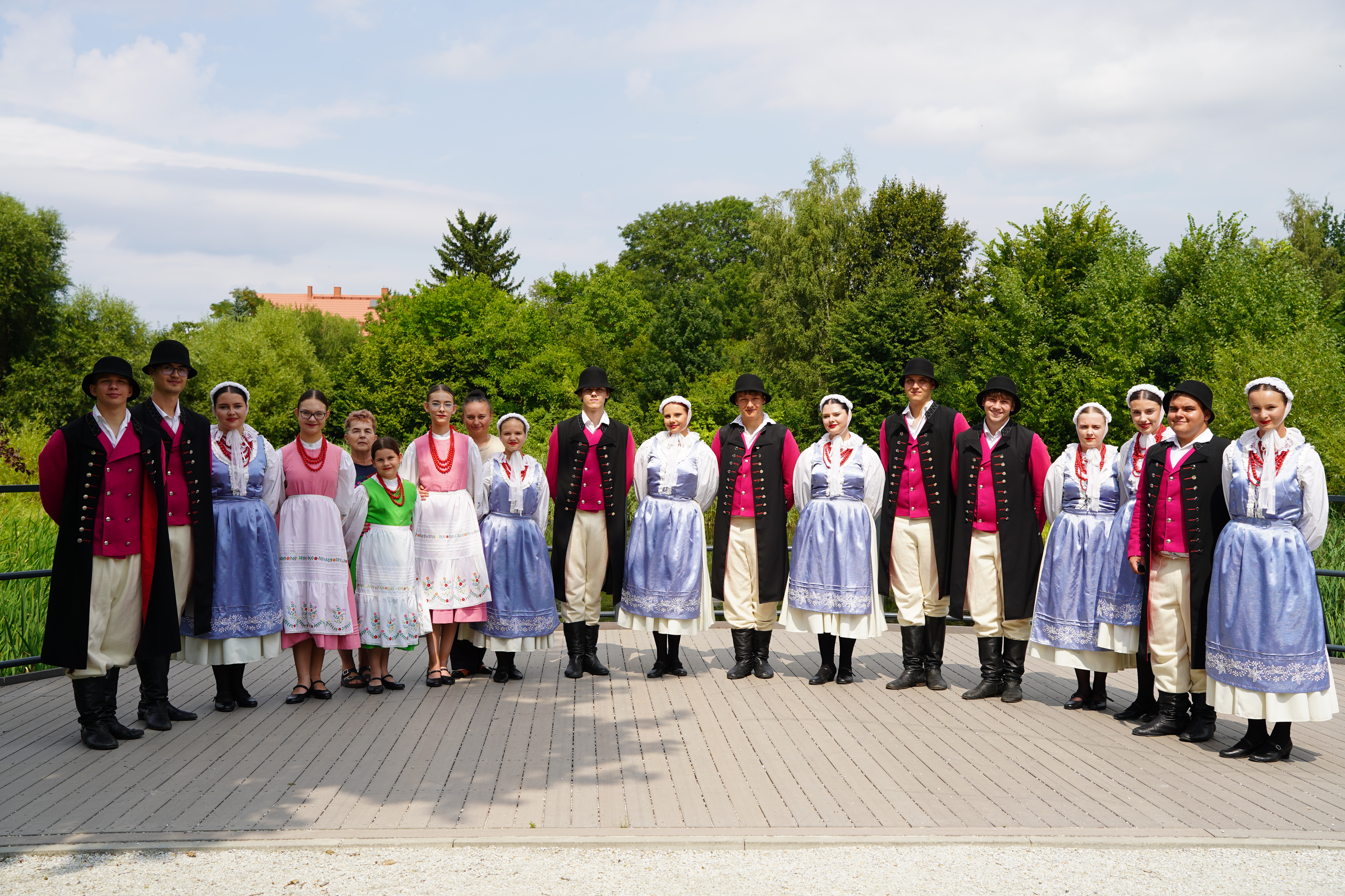 Zespół folklorystyczny, siedemnaście osób, pozuje w rzędzie na drewnianym pomoście. Mężczyźni w czarnych kapeluszach, długich płaszczach, amarantowych kamizelkach i wysokich butach; kobiety w błękitnych spódnicach i koralach. W tle zieleń drzew.