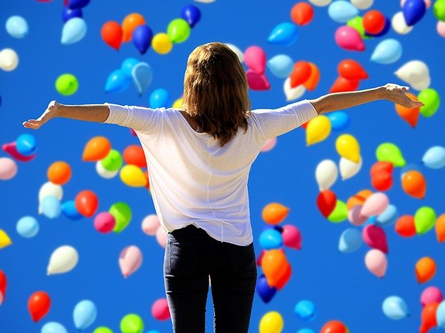 Zdjęcie przedstawia kobietę z rozpostartymi ramionami, w tle wypuszczone kolorowe balony.
