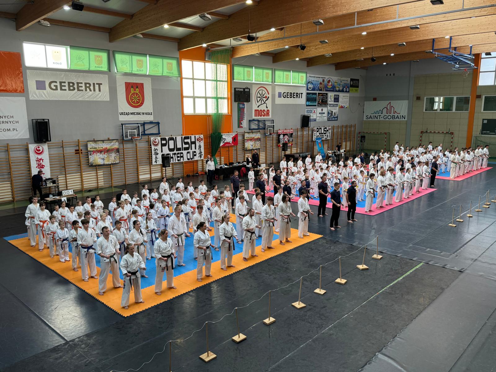 Na zdjęciu, wykonanym z trybun, widać zawodników w strojach do karate stojących na hali sportowej. W tle znajdują się logotypy różnych firm, duże okna oraz konstrukcja dachu obiektu.