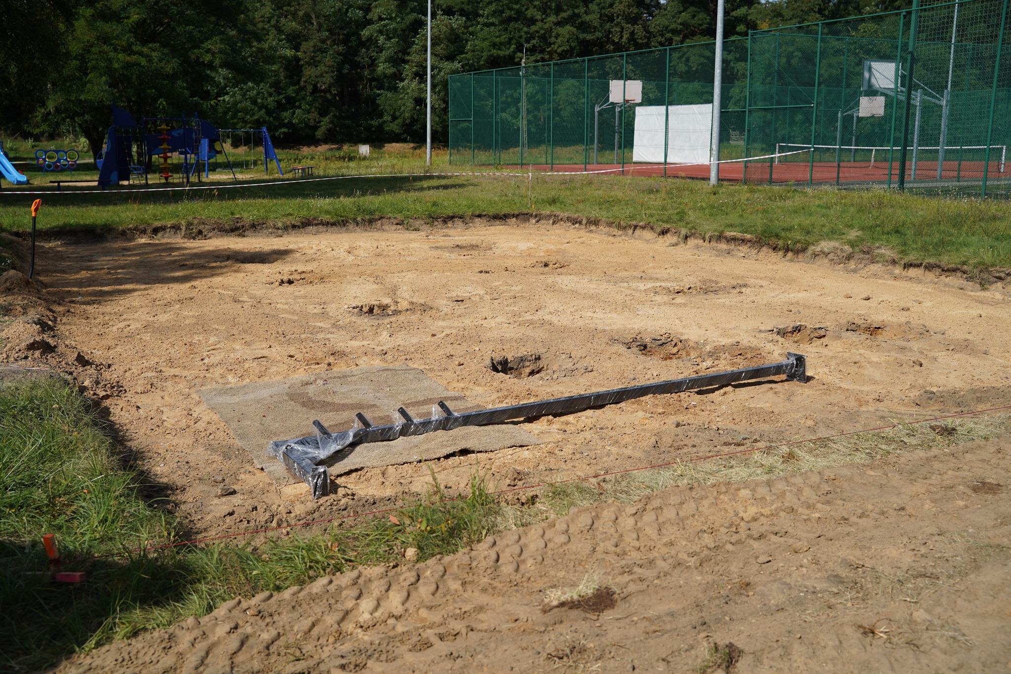 Na zdjęciu widać plac budowy na świeżo wyrównanym terenie z elementami fundamentów, a w tle znajduje się plac zabaw i boisko sportowe.