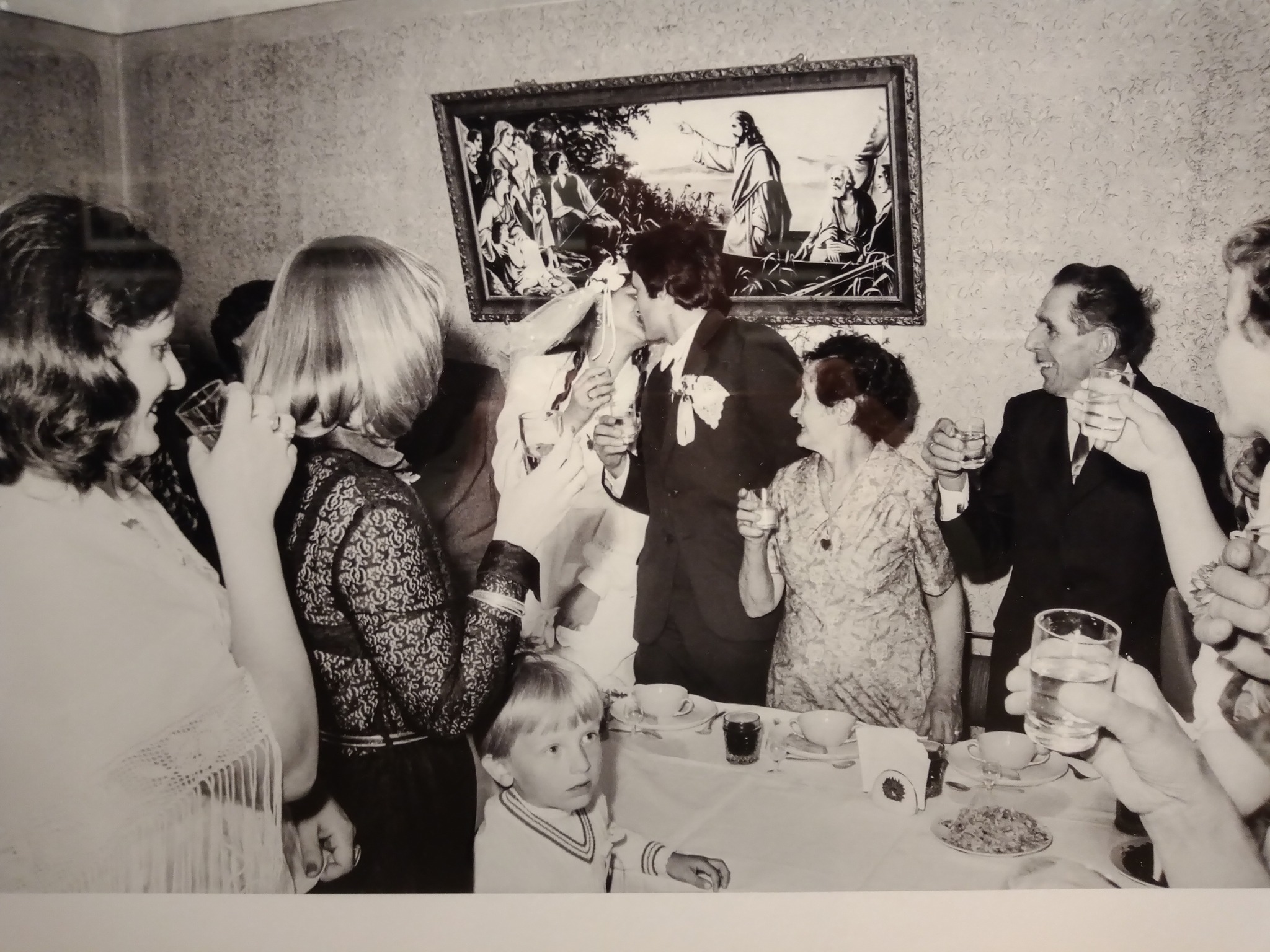Na czarno-białym zdjęciu grupa ludzi świętuje przy stole. W centrum młoda para całuje się, trzymając kieliszki, wokół goście wznoszą toasty. Na ścianie wisi obraz religijny, a na stole stoją filiżanki i przekąski. Wśród dorosłych widać małe dziecko.