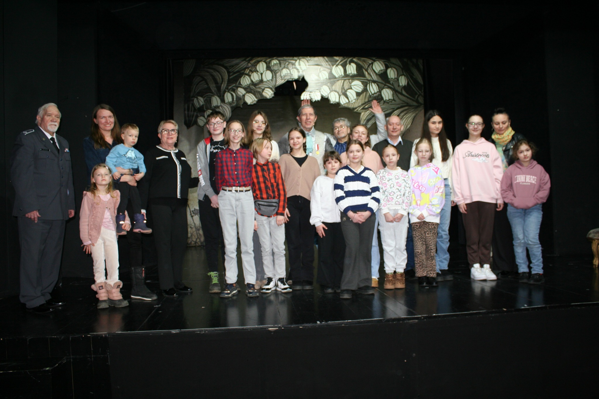 Na scenie teatru, na tle czarnej scenografii i dekoracji scenicznej, pozuje wspólnie grupa uśmiechniętych dzieci, młodzieży oraz opiekunów w towarzystwie aktorów, tworząc wyjątkową, pełną radości pamiątkę ze spotkania ze sztuką.