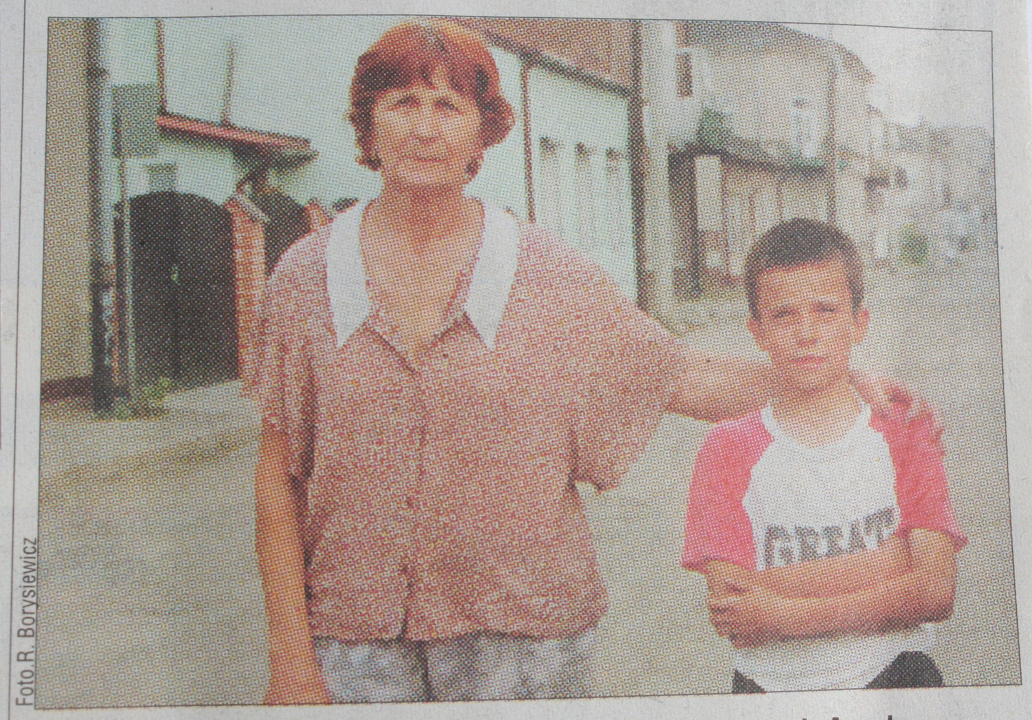 Archiwalne zdjęcie przedstawia kobietę i chłopca stojących na drodze. Kobieta trzyma lewą dłoń na ramieniu chłopca. Chłopiec ubrany jest w koszulkę z krótkim rękawem. W tle znajdują się budynki i stare kamienice.