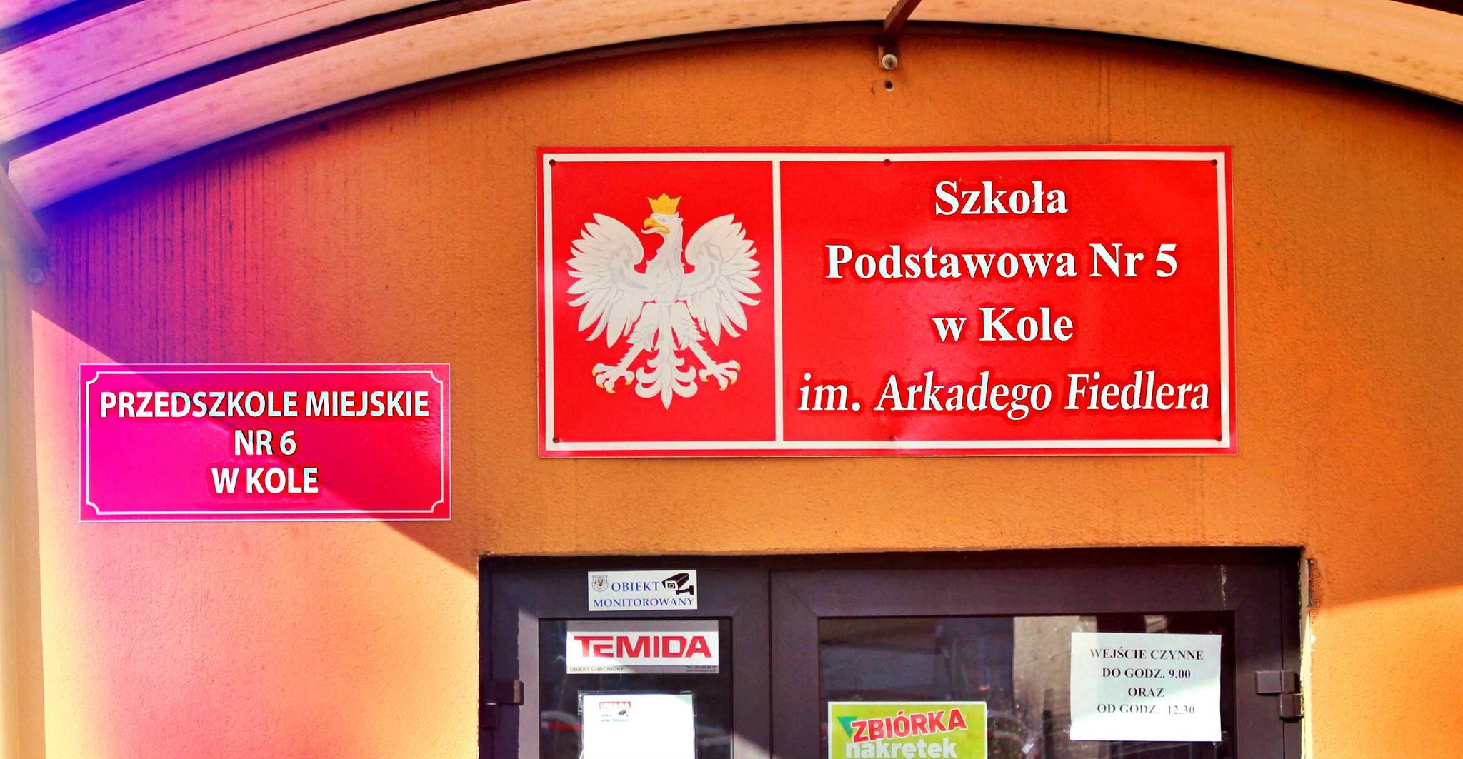 Zdjęcie przedstawia czerwone tablice z nazwą placówek nad wejściem do budynku szkoły i przedszkola