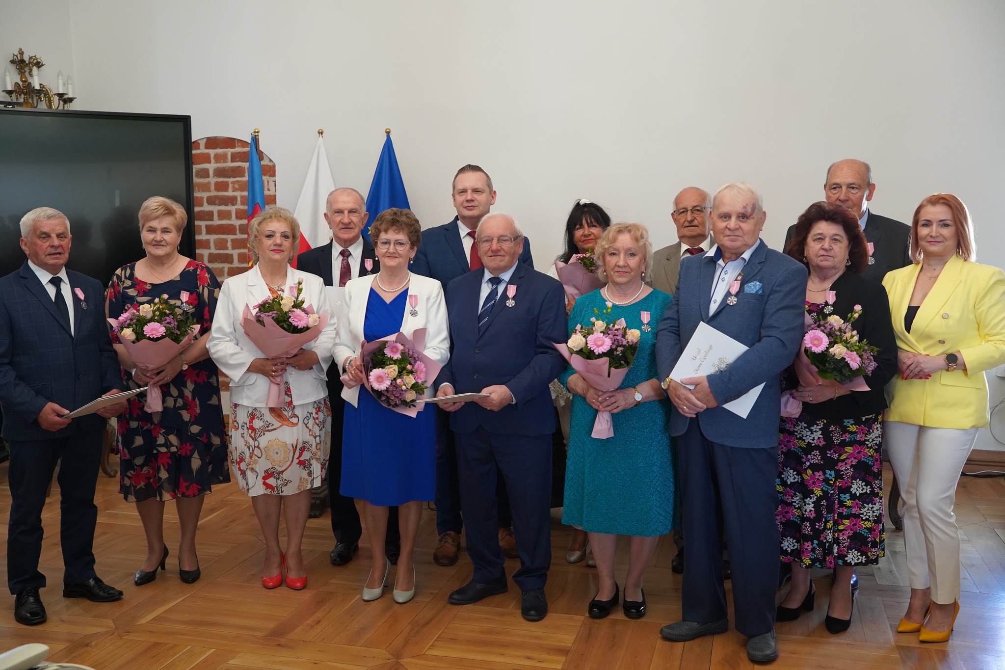 Na zdjęciu widać grupę elegancko ubranych osób, które trzymają odznaczenia i kwiaty z okazji jubileuszu pożycia małżeńskiego.