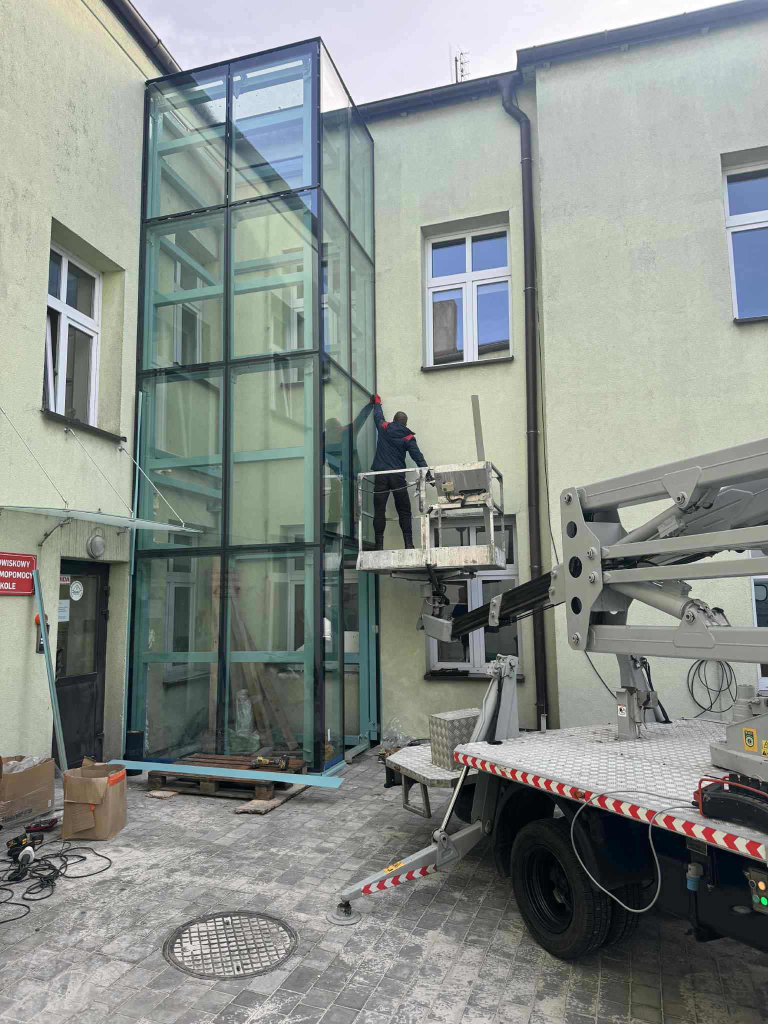 Na zdjęciu widać budynek gdzie trwają prace budowlane związane z montażem windy zewnętrznej. Na pierwszym planie widać zieloną, oszkloną, stalową konstrukcję, zamontowaną od parteru po ostatnie piętro budynku.