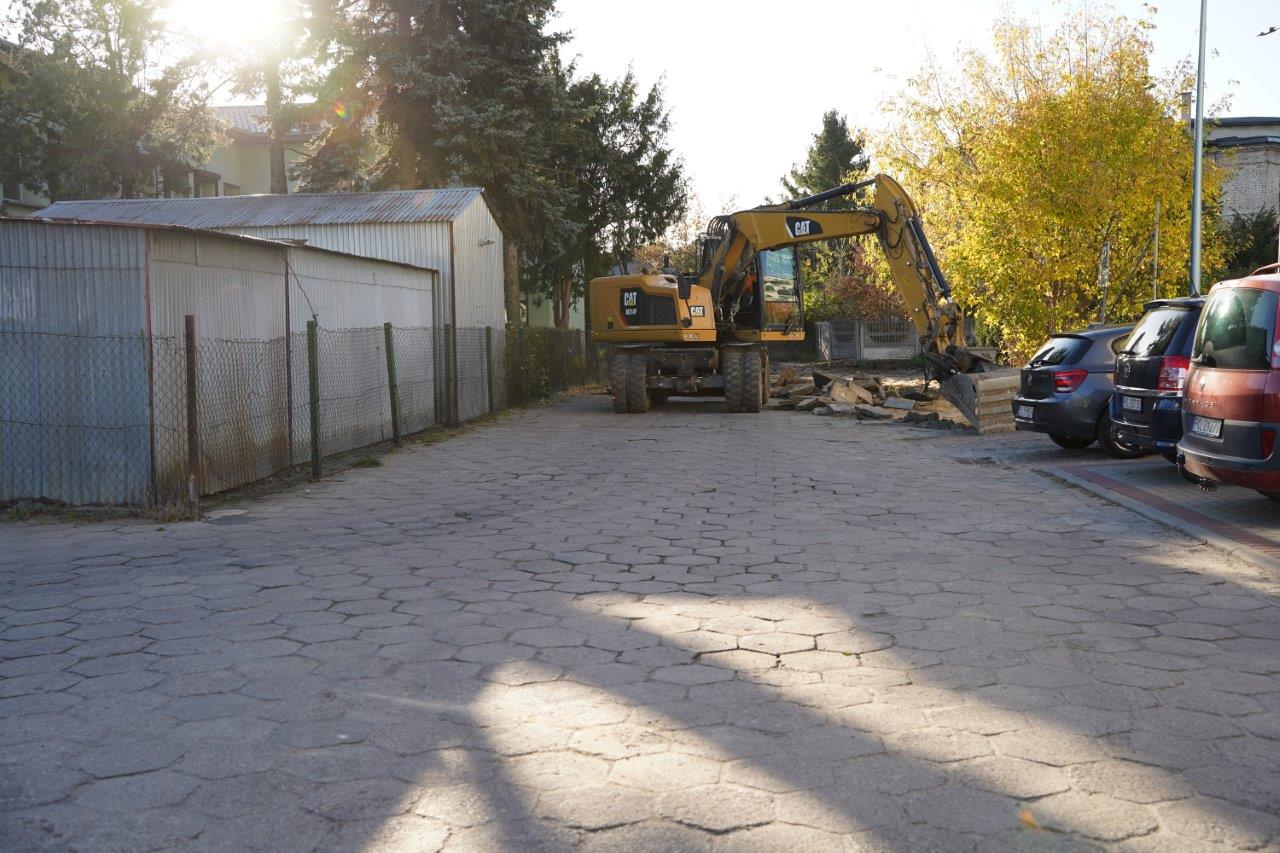 Osiedlowa droga z wyblakłej kostki sześciokątnej w jesiennym słońcu. W głębi żółta koparka kołowa CAT rozbiera nawierzchnię, obok sterta płyt betonowych. Po lewej blaszane garaże za siatką, po prawej zaparkowane auta i drzewa w żółtych liściach.