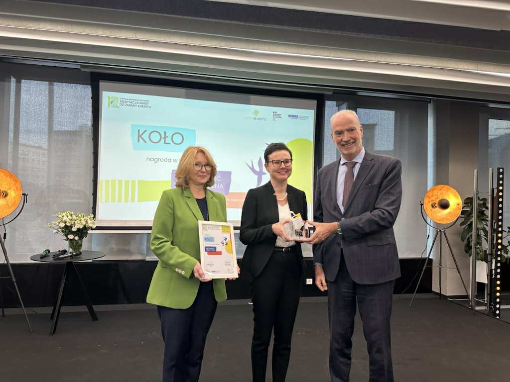 Na zdjęciu widać trzy osoby podczas uroczystości wręczenia nagrody dla miasta Koło Eco-Miasta 2025, odbywającej się na scenie z prezentacją w tle. Nagrodę odbiera Zastępca Burmistrza Monika Ciesielska.