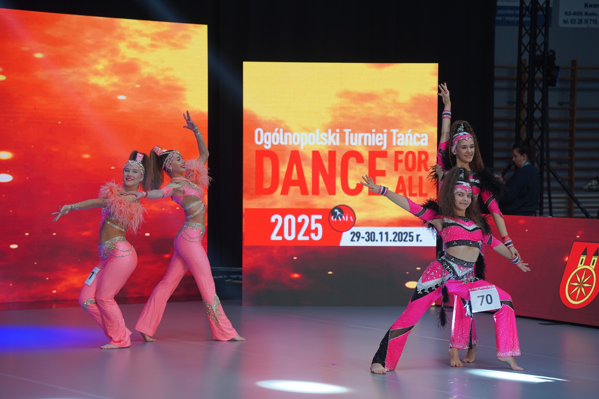 Na zdjęciu widać grupę młodych osób tańczących na scenie, ubranych w stroje taneczne. Tło stanowią telebimy, a oświetlenie sceniczne skupia się na tancerzach, podkreślając dynamiczny charakter ich ruchów.