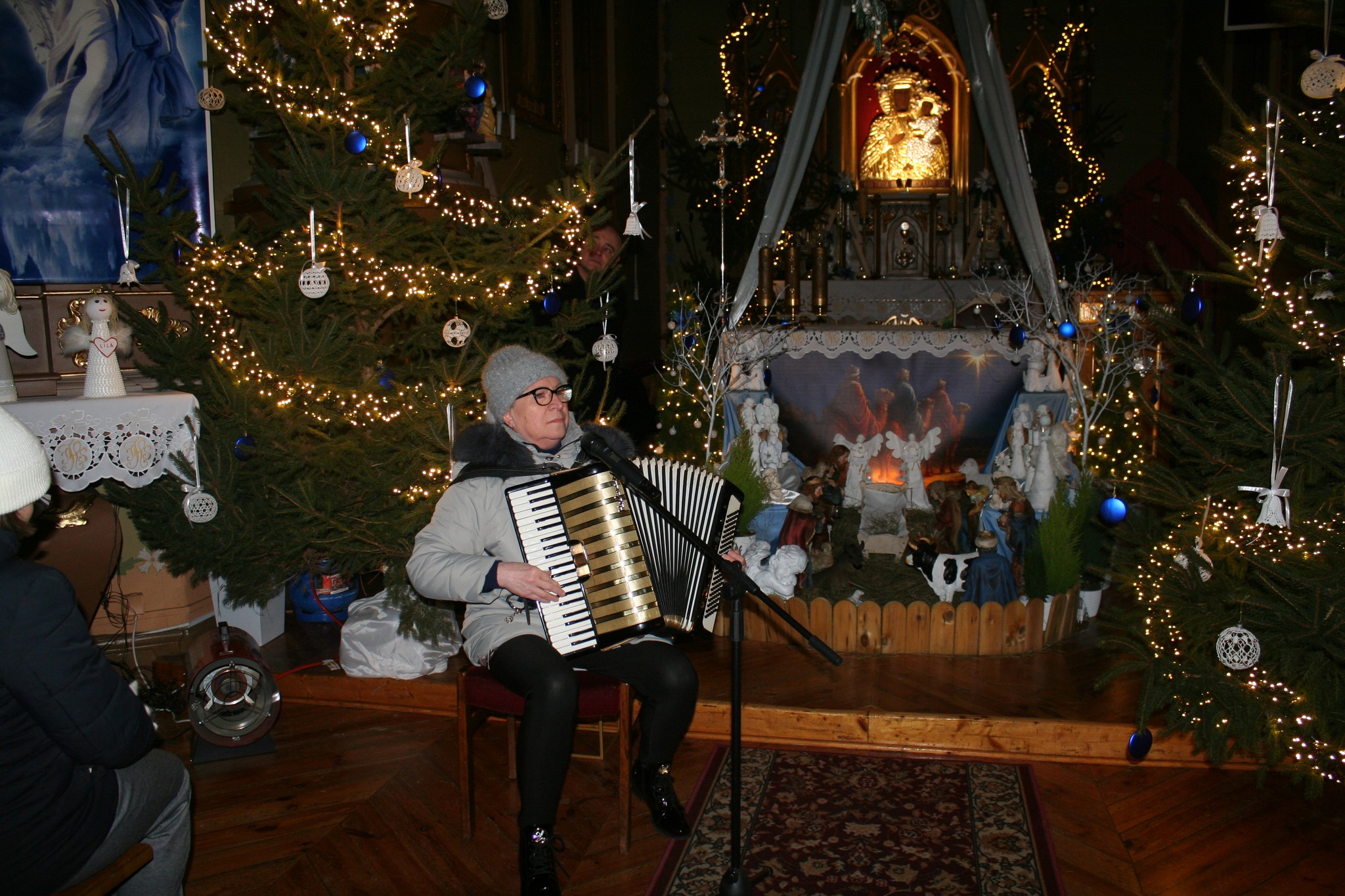 Na zdjęciu widać kobietę w kurtce i czapce, siedzącą na krześle i grającą na akordeonie w kościele. W tle znajduje się bożonarodzeniowy żłóbek, otoczony udekorowanymi żywymi choinkami.