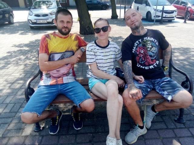 Na zdjęciu widzimy trzy osoby siedzące obok siebie na ławeczce na Starym Rynku przy Ratuszu w Kole. Dwie osoby są mężczyznami, a jedna jest kobietą. W tle zaparkowane samochody i pojedyncze drzewa.