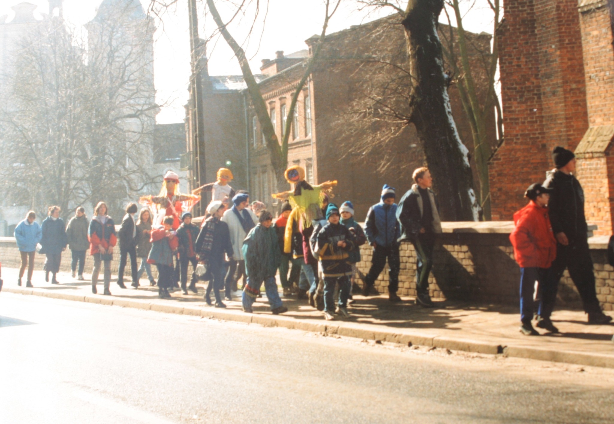 Grupa dzieci i młodzieży wraz z opiekunami maszeruje chodnikiem w kolorowych kurtkach, niosąc trzy marzanny; w tle widoczne są charakterystyczne budynki miasta – kościół farny, magistrat oraz klasztor.