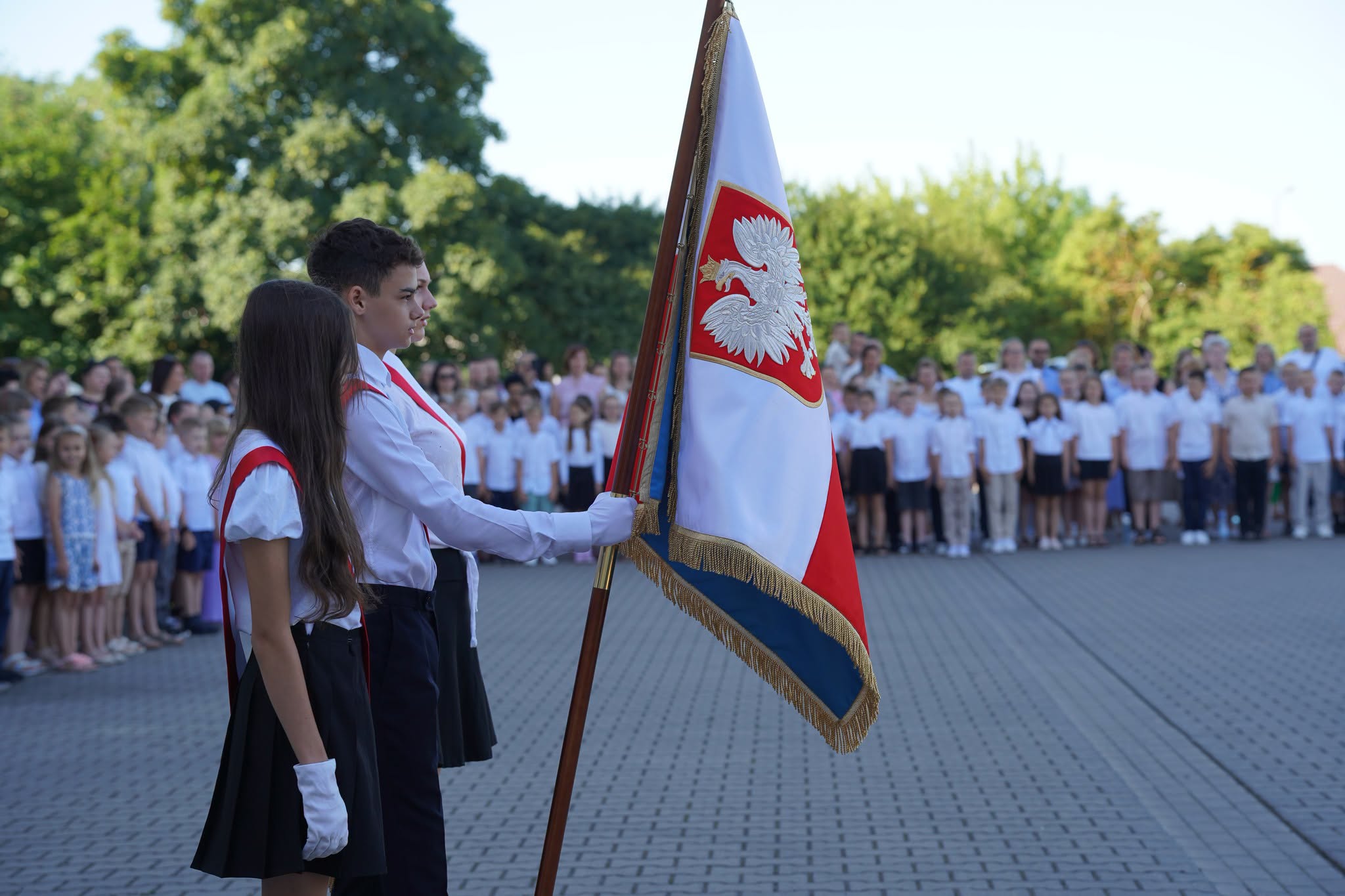 Uroczystość szkolna na placu. Dwoje uczniów w strojach galowych i białych rękawiczkach trzyma sztandar szkoły z godłem Polski. W tle zgromadzona społeczność szkolna w białych koszulach.