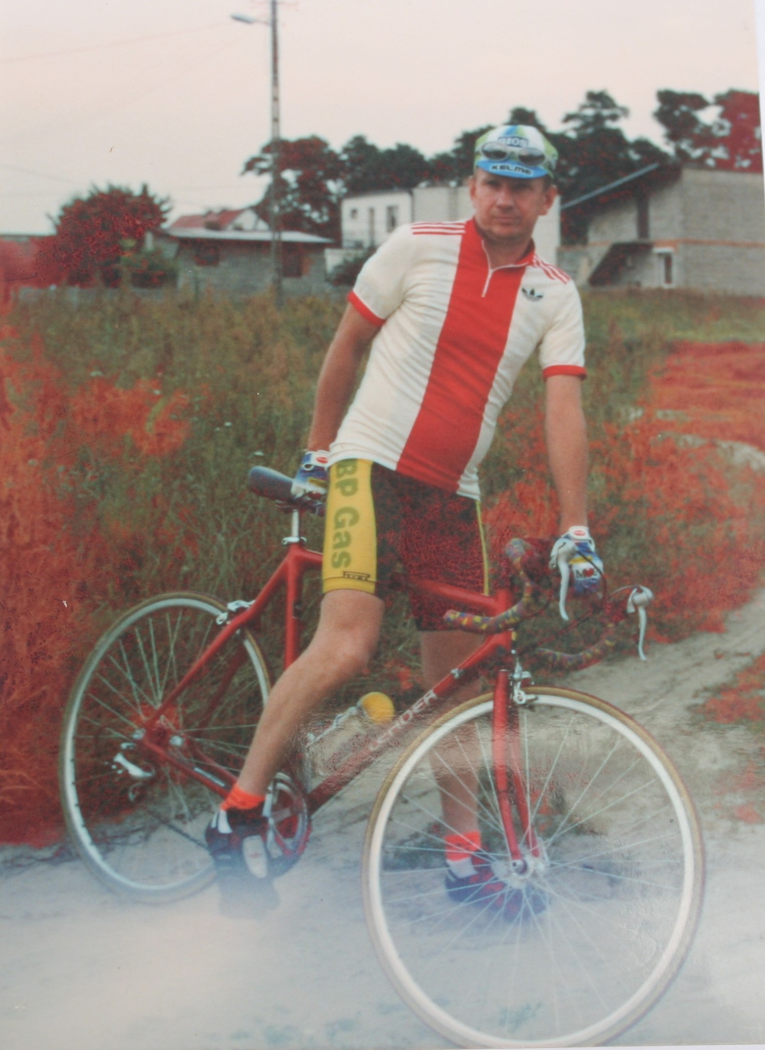 Zdjęcie przedstawia kolarza w stroju sportowym z rowerem szosowym, stojącego na nieutwardzonej drodze wśród traw i domów jednorodzinnych w tle. Styl ubioru i jakość fotografii sugerują, że pochodzi z lat 90.