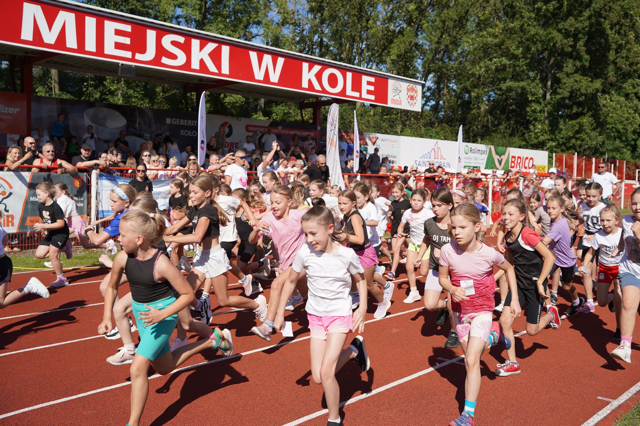 Zdjęcie przedstawia grupę małych dzieci biegnących na bieżni wokół stadionu. W tle widać rodziców przy ogrodzeniu. Pogoda jest słoneczna, a dzieci uczestniczą w zawodach sportowych w ramach 45. Międzynarodowego Biegu Warciańskiego 
