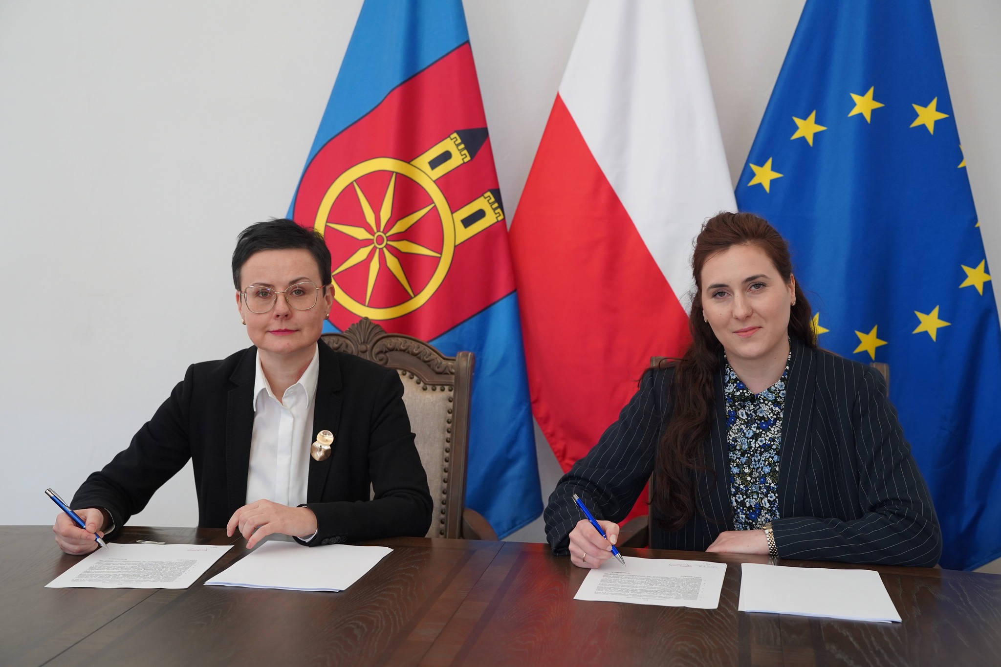 Na zdjęciu widać dwie kobiety siedzące przy stole prezydialnym w sali sesyjnej Ratusza w Kole, które podpisują umowę. W tle widać trzy flagi: miasta Koła, Polski oraz Unii Europejskiej. 