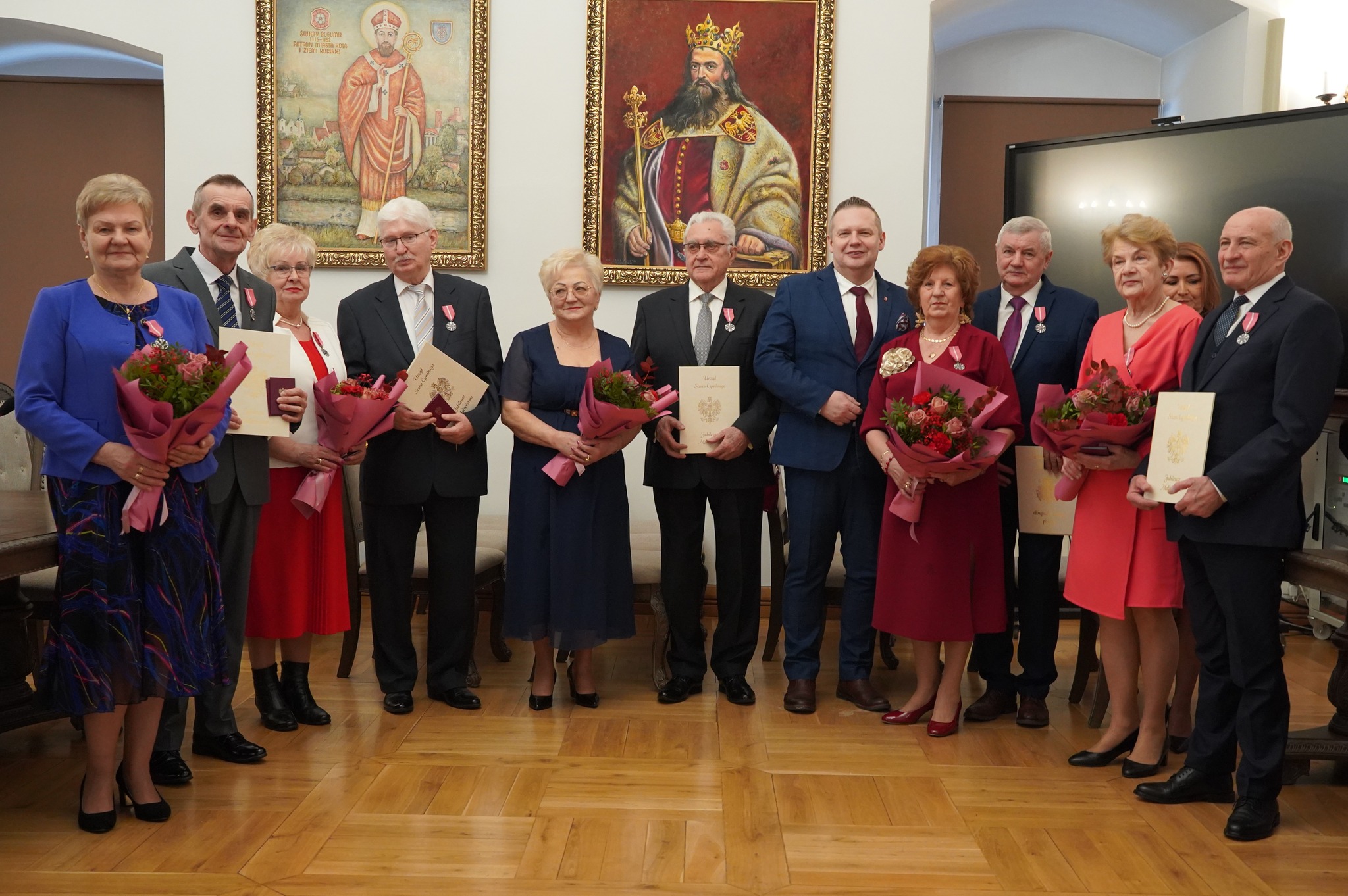 Na zdjęciu widać grupę elegancko ubranych osób, które trzymają odznaczenia i kwiaty z okazji jubileuszu pożycia małżeńskiego. W tle na ścianie widać wiszące obrazy króla Kazimierza Wielkiego i św. Bogumiła. 