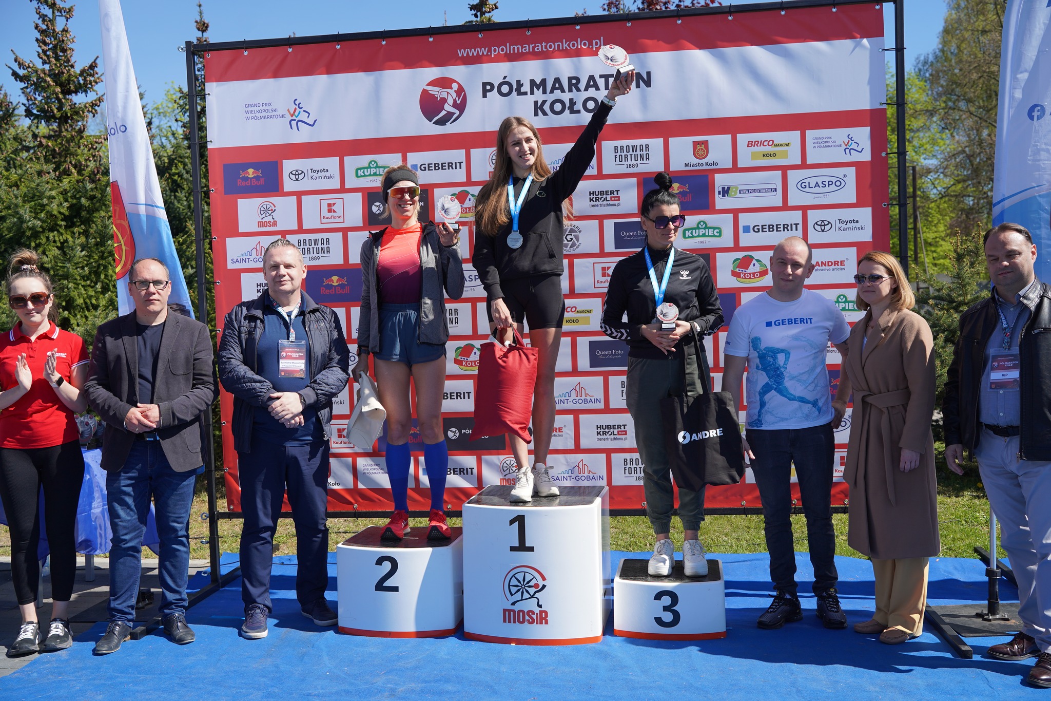 Na zdjęciu widać podium z trzema kobietami – zwyciężczyni stoi na najwyższym stopniu i unosi medal. Po bokach podium stoją organizatorzy i goście wydarzenia. W tle widoczna jest ścianka z logotypami sponsorów i napisem Półmaraton Koło
