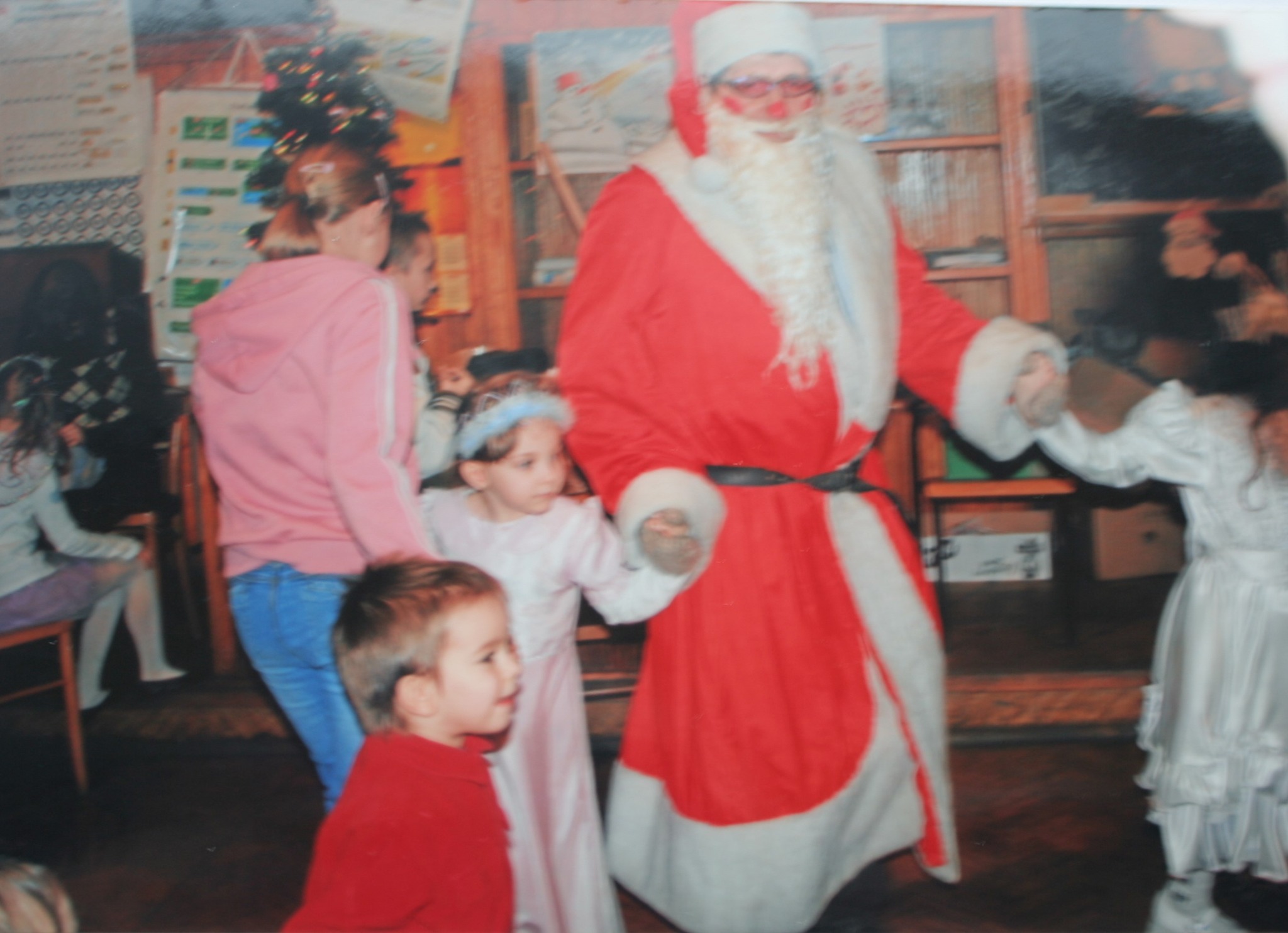 Na zdjęciu widać osobę przebraną za Świętego Mikołaja, która trzyma za ręce dzieci podczas świątecznej zabawy w sali, wśród choinki i innych uczestników.