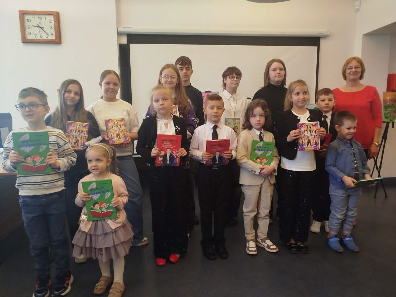 Grupa dzieci – dziewczynek i chłopców, niektóre trzymające dyplomy – pozuje do zdjęcia, obok nich stoi kobieta, a w tle widać ekran na ścianie, zegar oraz apteczkę.