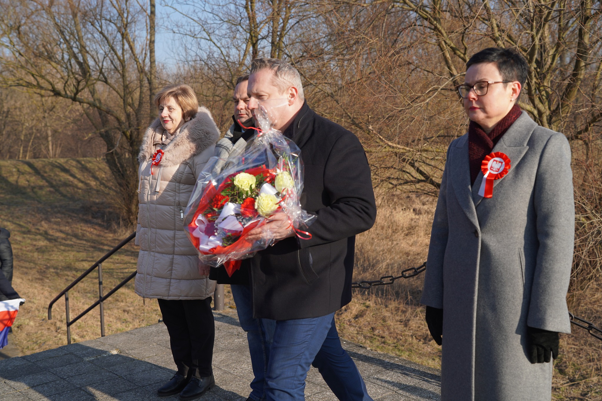 Na zdjęciu widać kilka osób stojących na zewnątrz podczas uroczystości patriotycznej. Jedna z osób trzyma bukiet kwiatów, a uczestnicy mają przypięte biało-czerwone kokardy. Scena wygląda na oficjalne składanie kwiatów w miejscu pamięci.