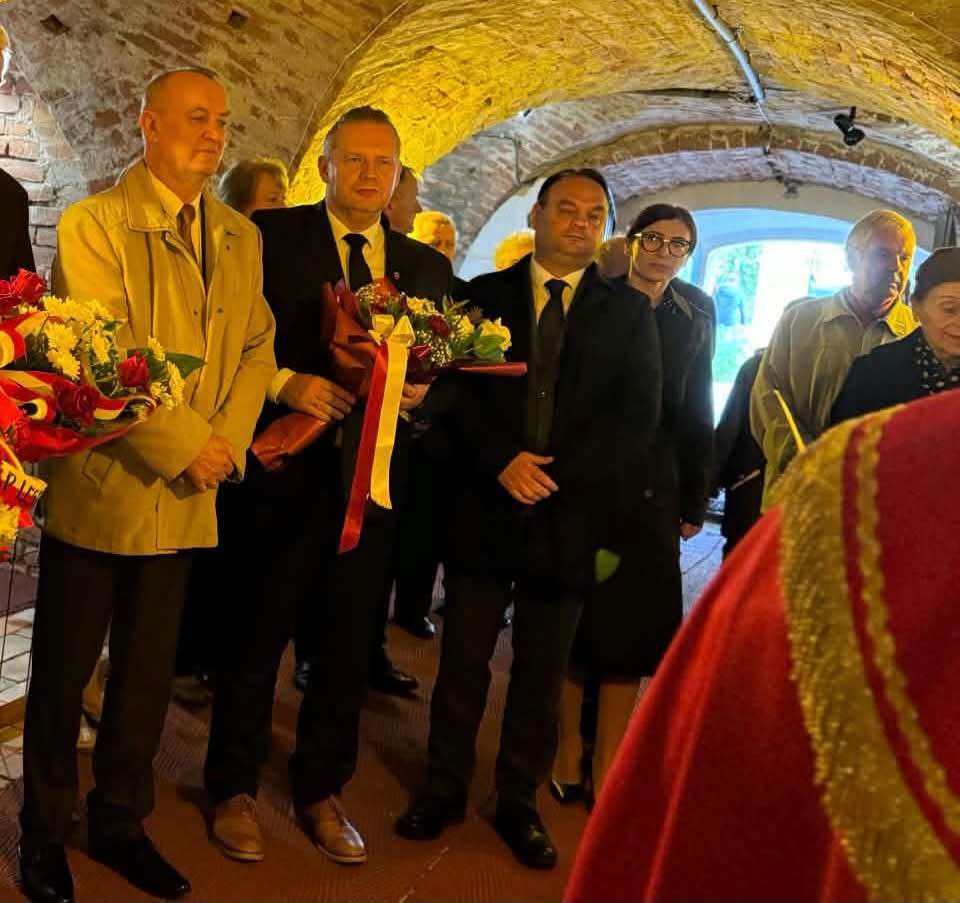 Uroczystość w podziemiach klasztoru z ceglanym sklepieniem. Kilka osób w ciemnych garniturach i jasnym płaszczu stoi w skupieniu. Trzymają wiązanki kwiatów z biało-czerwonymi szarfami. Za nimi są inni uczestnicy, na pierwszym planie czerwona szata. 
