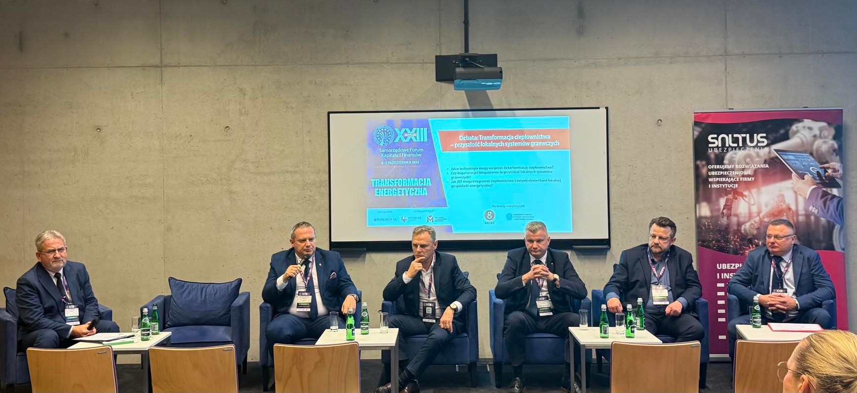 Na zdjęciu widać panel dyskusyjny z udziałem sześciu mężczyzn w garniturach, siedzących na scenie przed ekranem z prezentacją. Dyskusja dotyczy transformacji energetycznej i przyszłości lokalnych systemów grzewczych.
