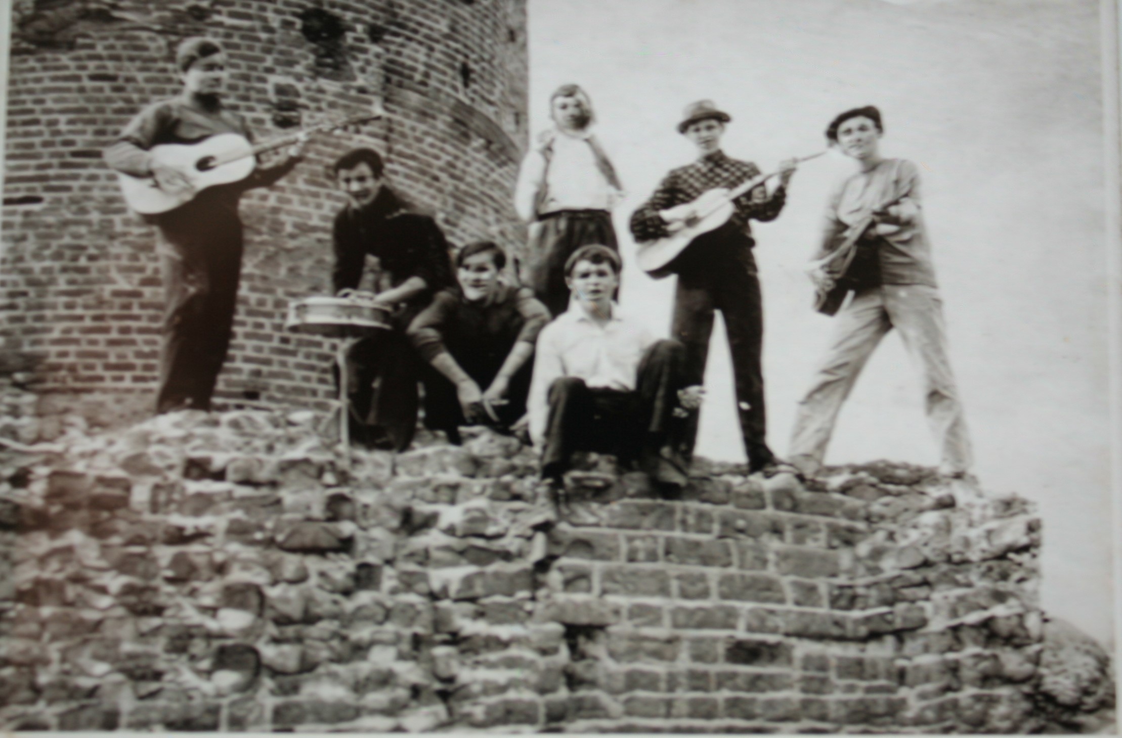 Na czarno-białym zdjęciu siedmioro nastolatków stoi na ruinach zamku, troje trzyma gitary, dwóch bęben i trąbkę, a w tle widać majestatyczną wieżę, tworząc atmosferę młodzieńczej pasji i muzycznej zabawy.