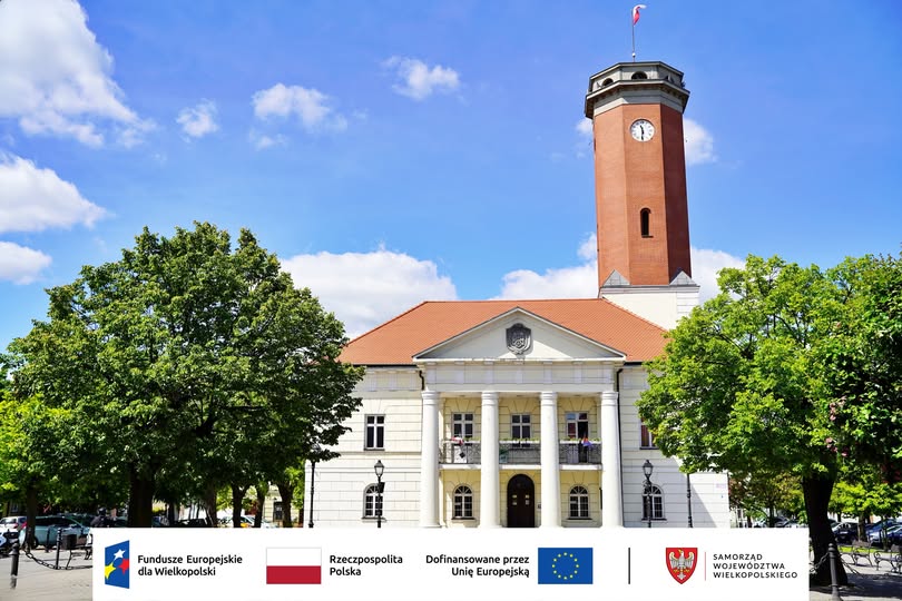 Na zdjęciu widać budynek ratusza z wieżą zegarową, otoczony drzewami, a na dole znajdują się logotypy Funduszy Europejskich, Rzeczypospolitej Polskiej, Unii Europejskiej i Samorządu Województwa Wielkopolskiego