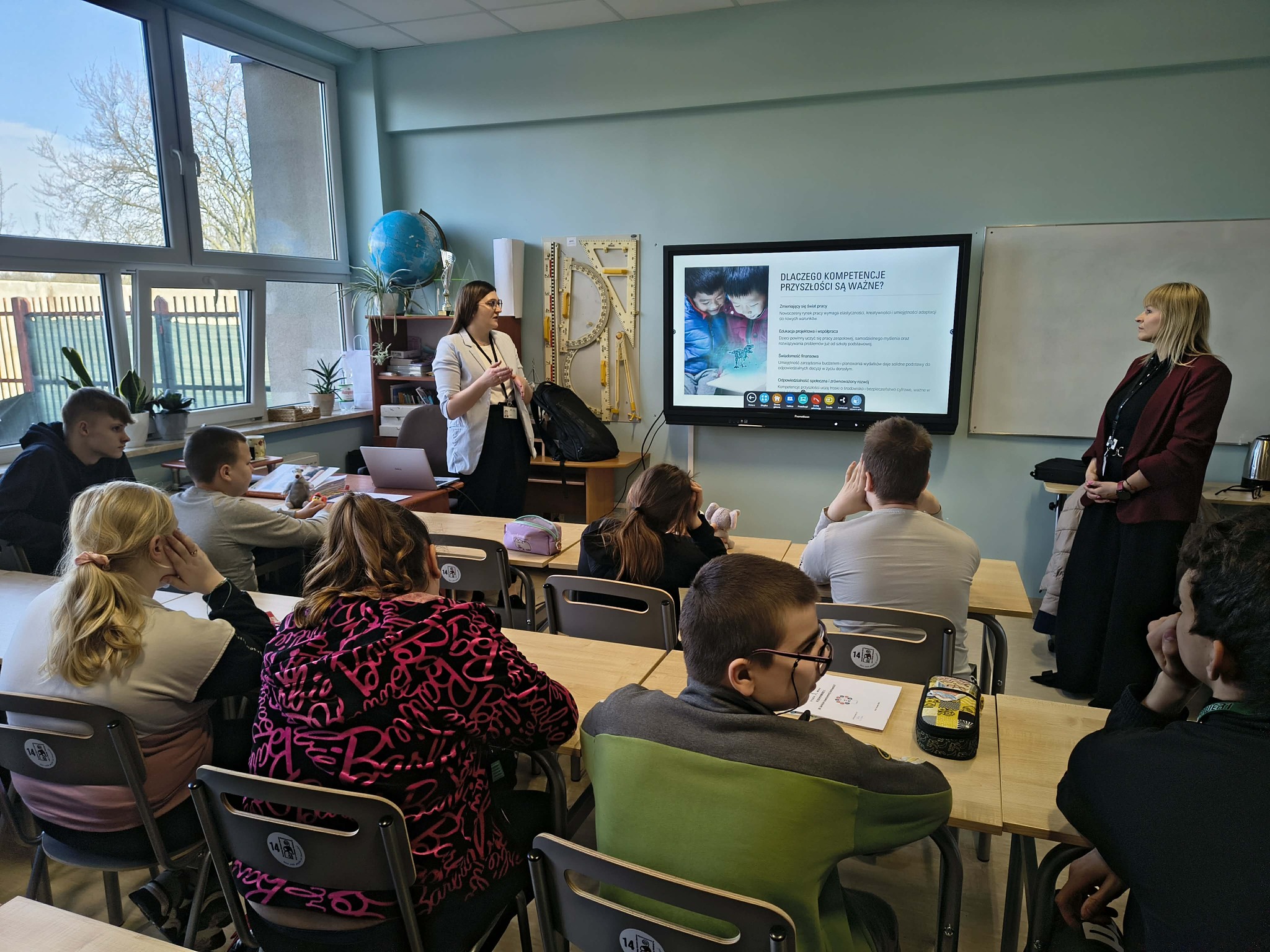 Prelegentka prowadzi wykład przed klasą, spoglądając na wiszący ekran z prezentacją, podczas gdy uczniowie siedzą w ławkach, a z prawej strony nauczycielka również patrzy w ekran.