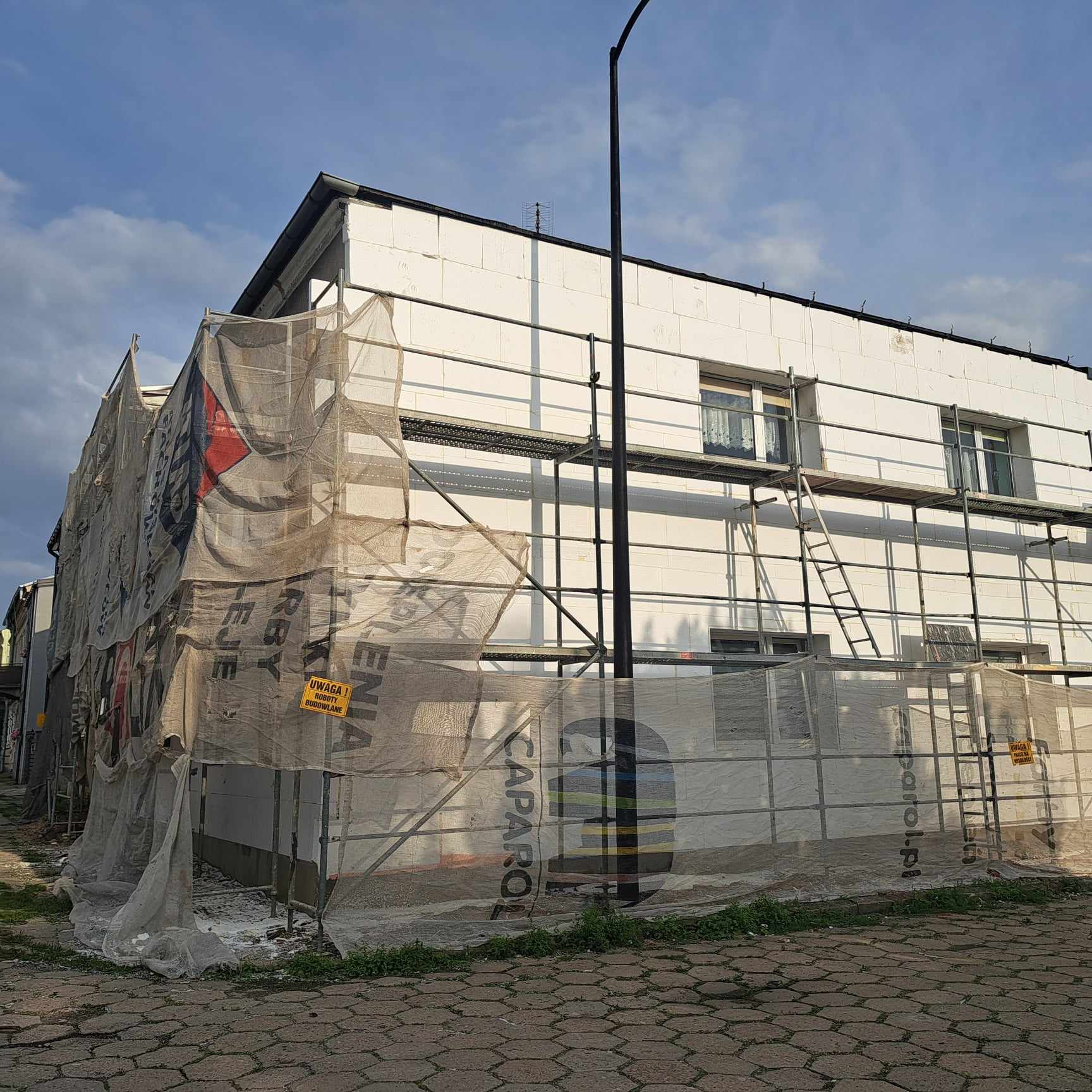 Na zdjęciu widzimy budynek, który jest w trakcie prac remontowych związanych z termomodernizacją. Na fasadzie widoczne są płyty izolacyjne, a wokół budynku znajduje się rusztowanie zabezpieczone siatkami ochronnymi. 
