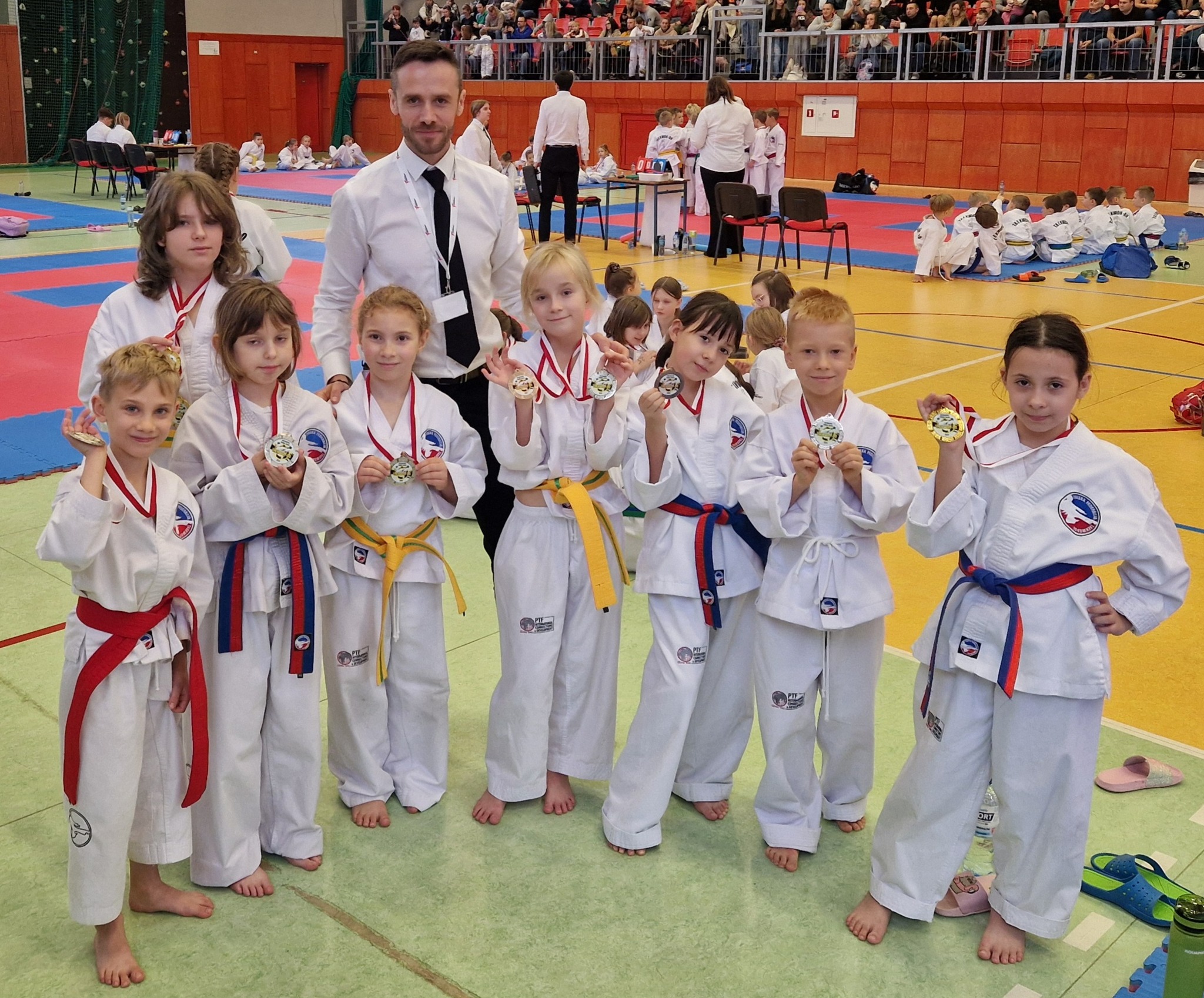 Grupa karateków, dziewcząt i chłopców wraz z trenerem, w hali sportowej, pozuje do zdjęcia z medalami na Mistrzostwach Warmii i Mazur Taekwon-do w Lidzbarku. W tle widać innych uczestników zawodów oraz kibiców na trybunach