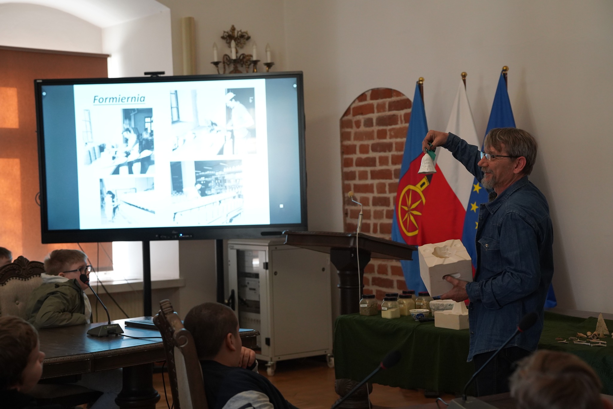 Na zdjęciu wykonanym w sali ratusza widać z lewej strony duży ekran z prezentacją oraz kilku słuchaczy siedzących na krzesłach, po prawej stoi prelegent, trzymający w prawej ręce ceramiczny dzwonek, a w lewej formę ceramiczną; w tle widoczne są flagi