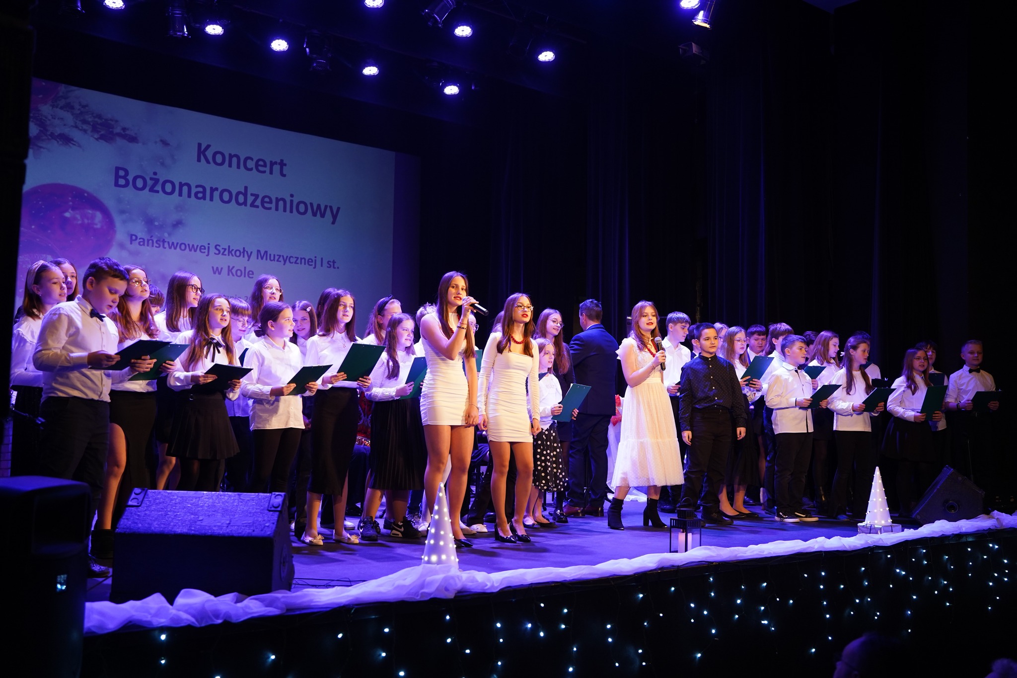 Na scenie występuje duża grupa dzieci i młodzieży ubranych w eleganckie czarno-białe stroje; na pierwszym planie, w centrum, widać trzy śpiewające dziewczyny, a w tle ekran z napisem Koncert Bożonarodzeniowy.