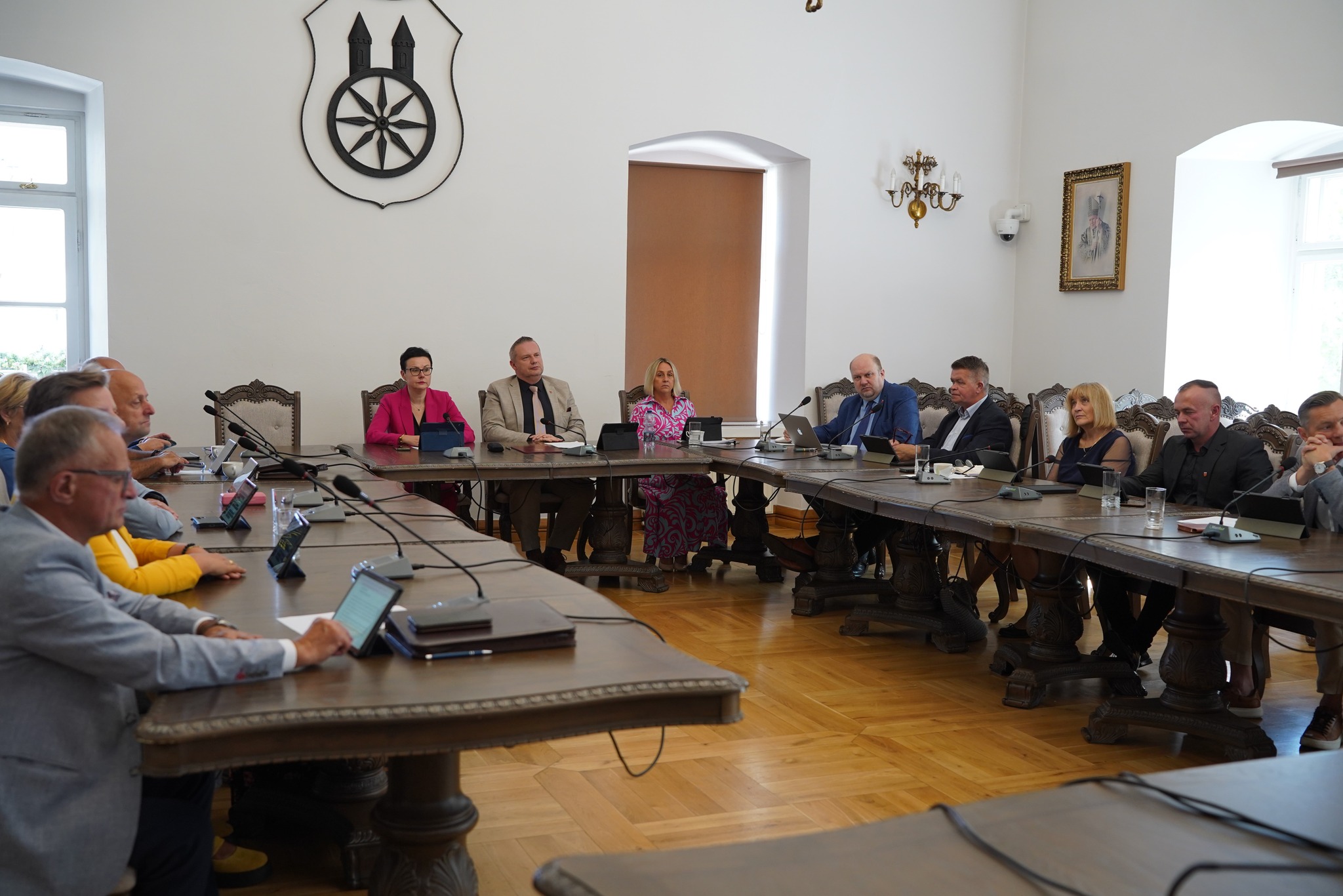Zdjęcie przedstawia obrady sesji rady miejskiej w Kole. Uczestnicy siedzą przy dużym, drewnianym stole, korzystając z tabletów i mikrofonów. W tle widoczne są herby oraz obrazy historycznych postaci. Sala ma elegancki, zabytkowy wystrój.
