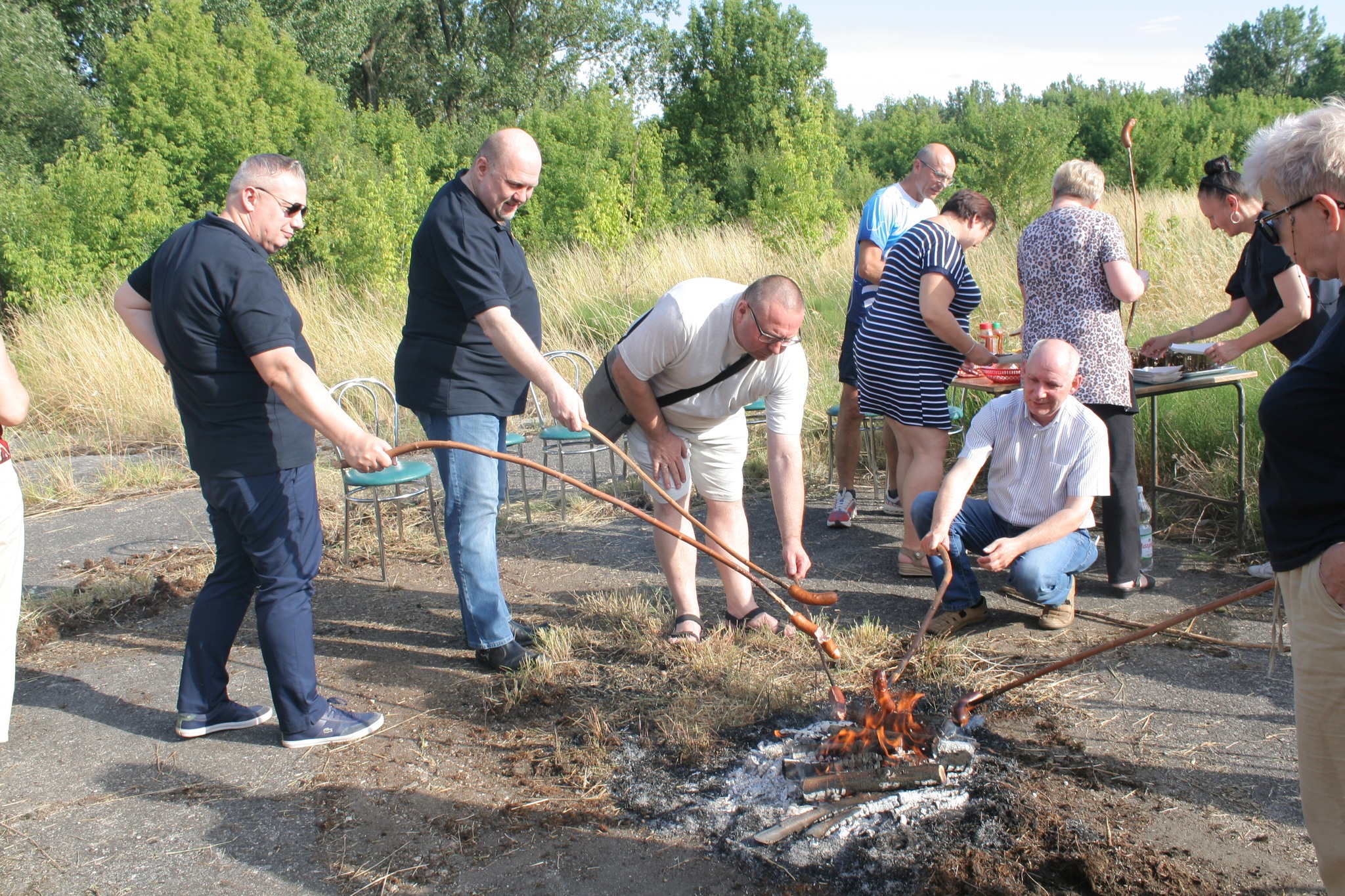 Na zdjęciu widać grupę osób piekących kiełbaski nad ogniskiem podczas plenerowego spotkania, z okazji rozpoczęcia wakacji.