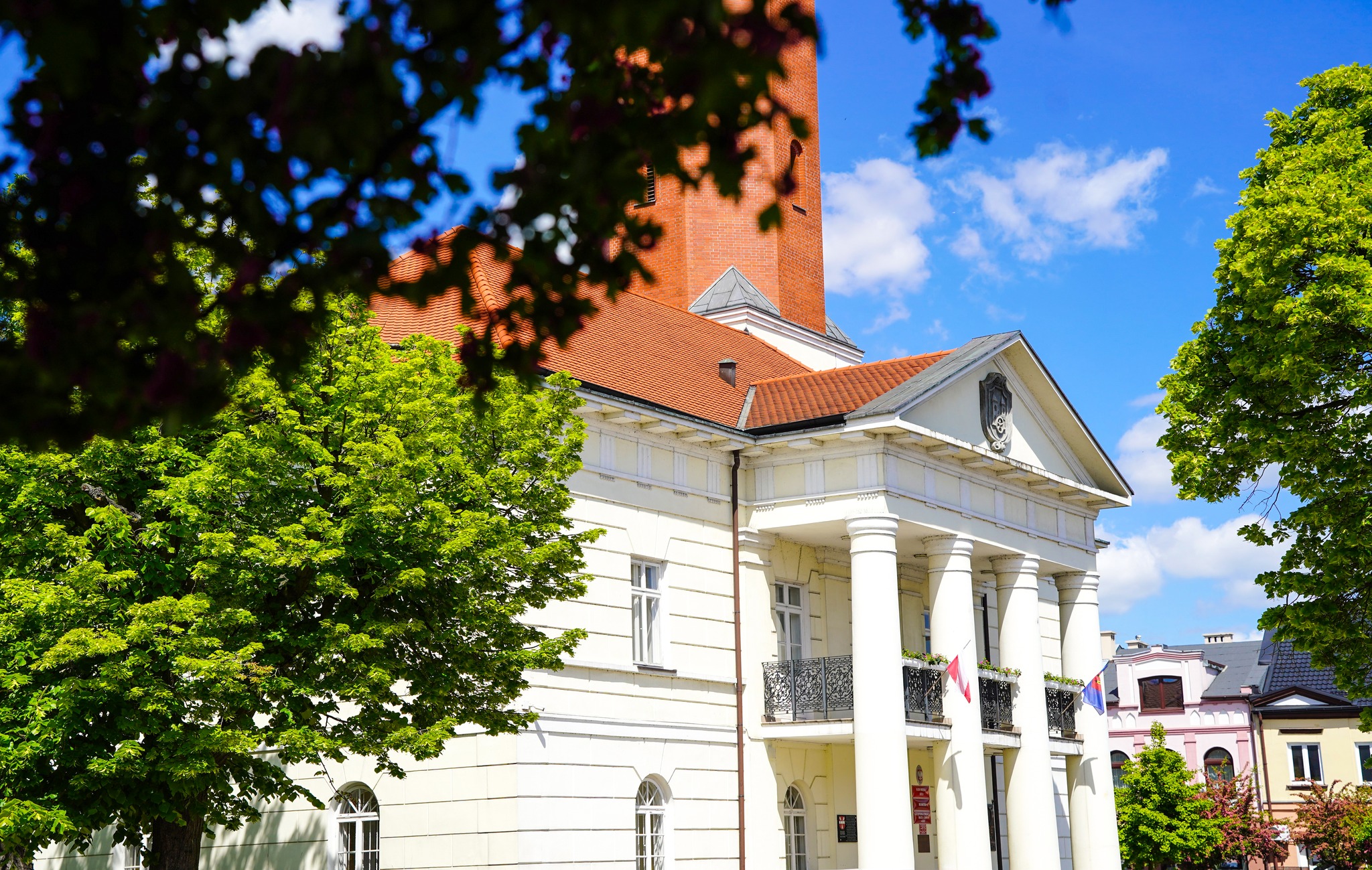 Zdjęcie przedstawia ratusz w mieście Koło. Budynek ma klasyczny wygląd, z charakterystycznymi białymi kolumnami na fasadzie, które wspierają balkon nad wejściem. Widać również czerwoną wieżę zegarowa z polską flagą na szczycie. Ratusz otaczają drzewa
