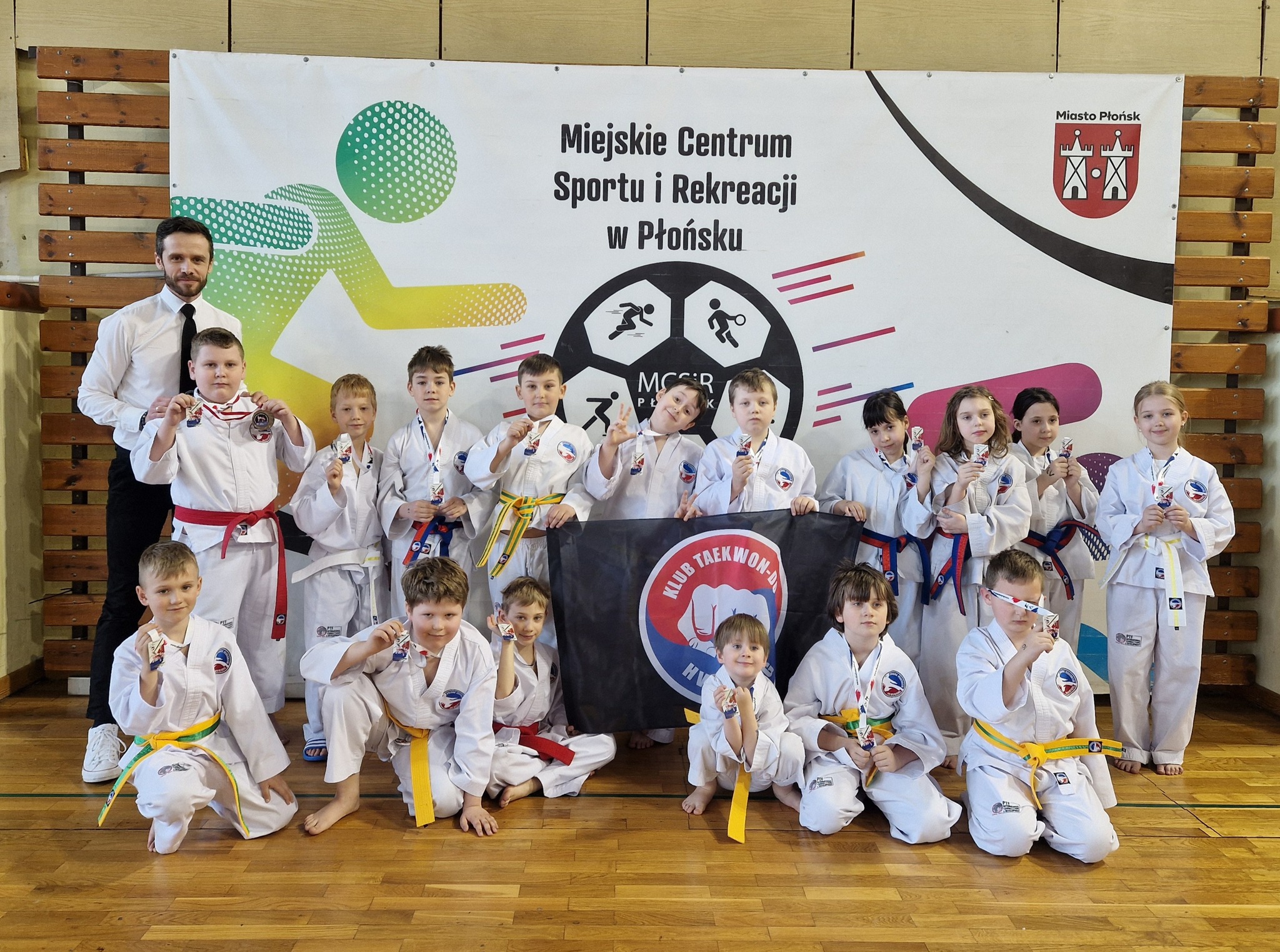 Zawodnicy Kolskiego Klubu Taekwon-do Hwarang pozują do wspólnego zdjęcia z medalami wraz z opiekunem. Zdjęcie zrobiono w hali sportowej. W tle duży baner powieszony na drabinkach z napisem Miejskie Centrum Sportu i Rekreacji w Płońsku.