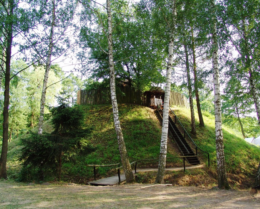 Na zdjęciu widać skansen – drewniane grodzisko otoczone palisadą, usytuowane na wzniesieniu wśród drzew, z prowadzącymi do niego schodami.