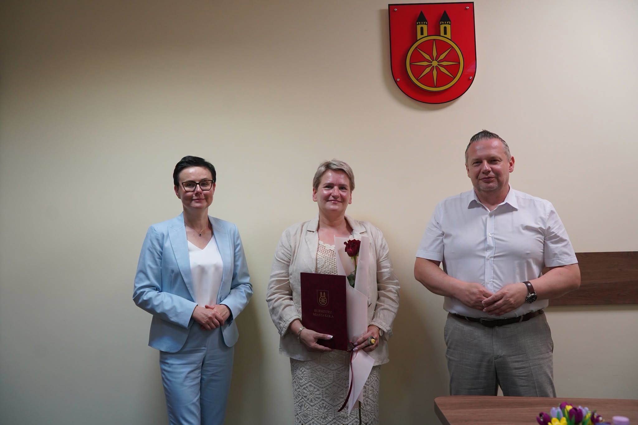 Trzy osoby stoją na tle ściany z herbem Miasta Koła. Kobieta w środku trzyma bordową teczkę z napisem Burmistrz Miasta Koła i czerwoną różę. Po lewej kobieta w błękitnym garniturze, po prawej mężczyzna w białej koszuli.