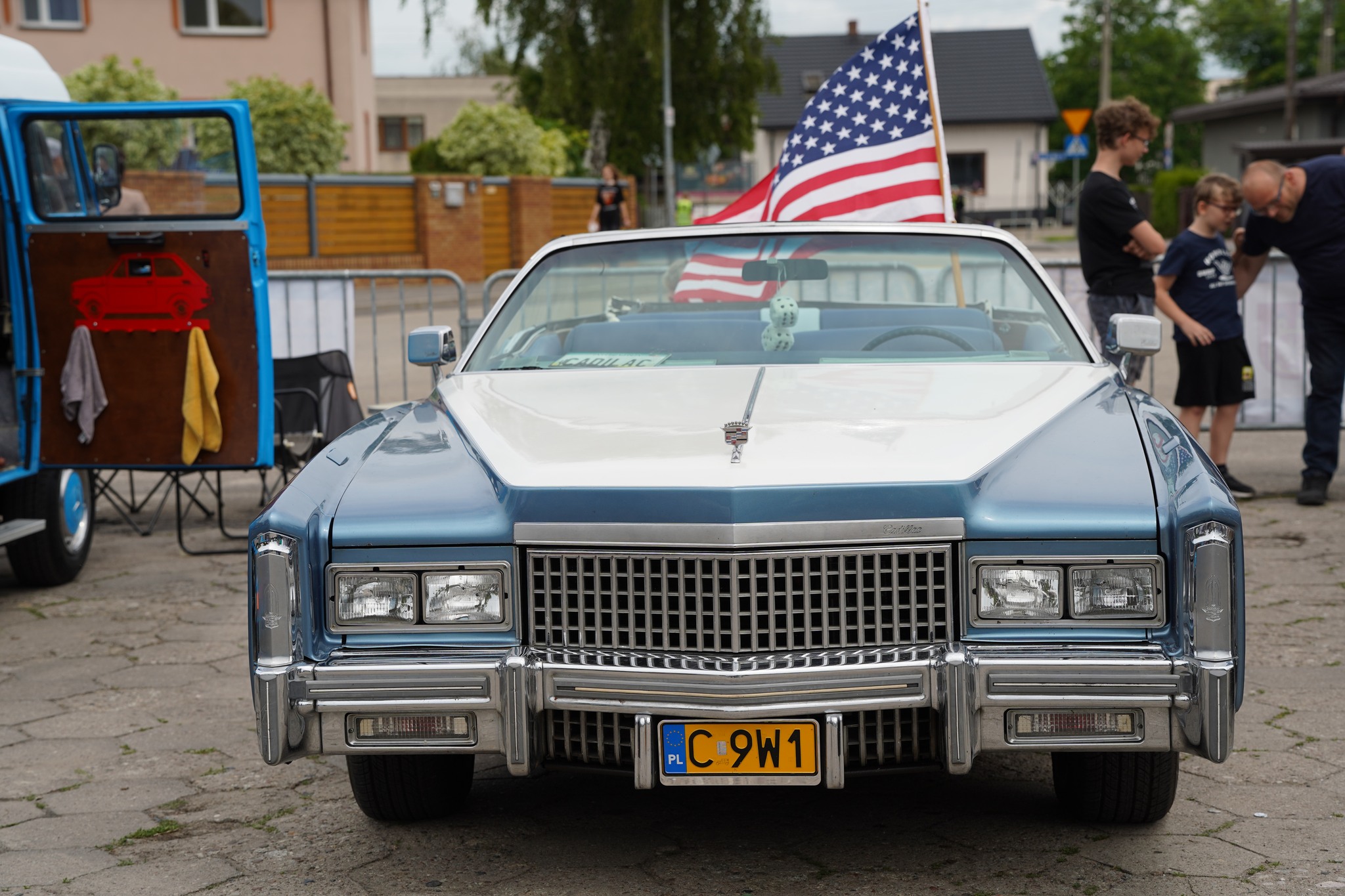 Na zdjęciu widać klasyczny amerykański samochód Cadillac z otwartym dachem, ozdobiony flagą USA. Auto stoi na zlocie pojazdów, w tle widać ludzi oraz inne pojazdy.