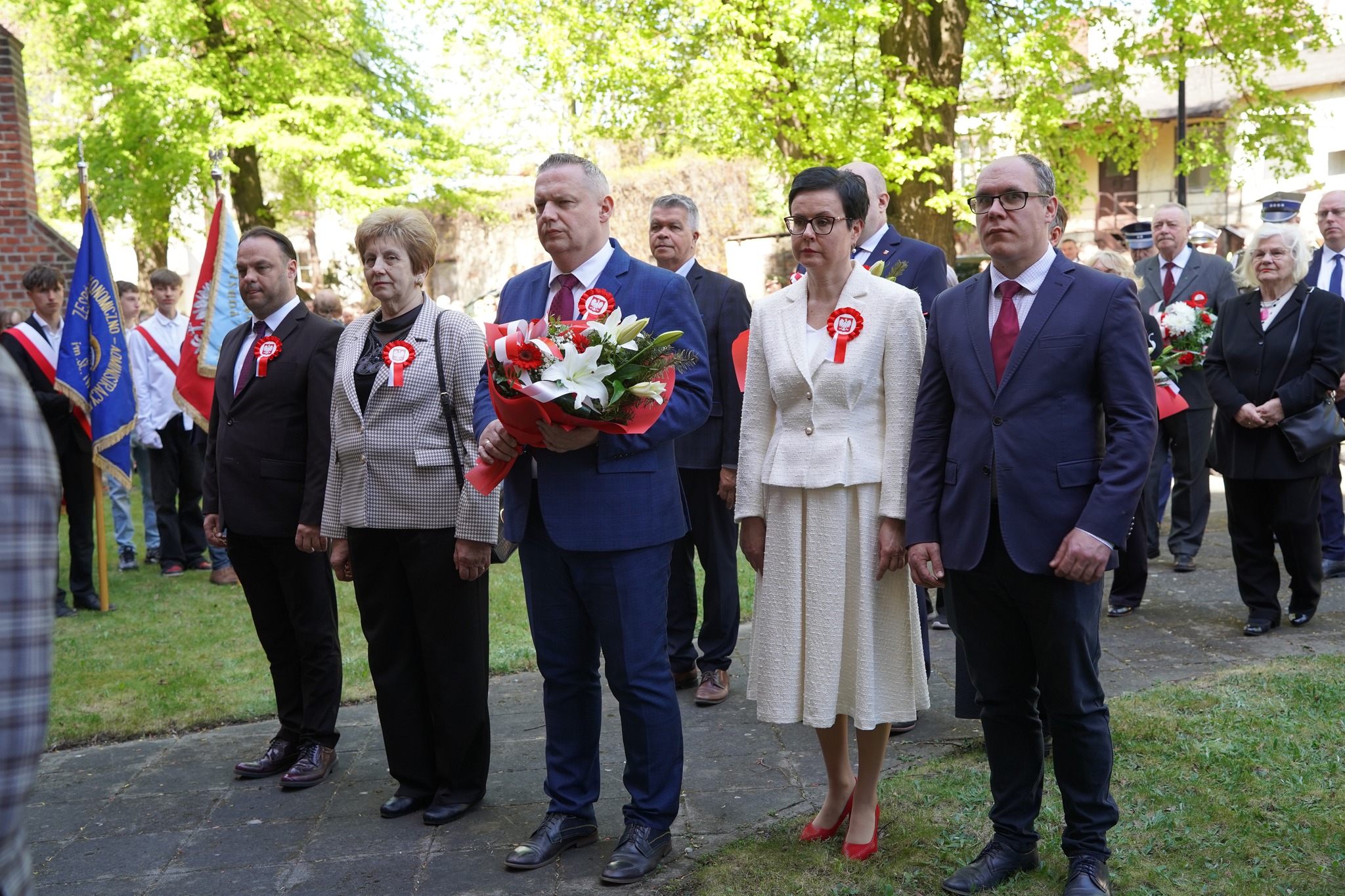 Na pierwszym planie widać pięcioosobową delegację składającą kwiaty: trzech mężczyzn i dwie kobiety, elegancko ubranych. Jeden z mężczyzn trzyma bukiet. W tle widoczne są drzewa oraz inne osoby uczestniczące w uroczystości.