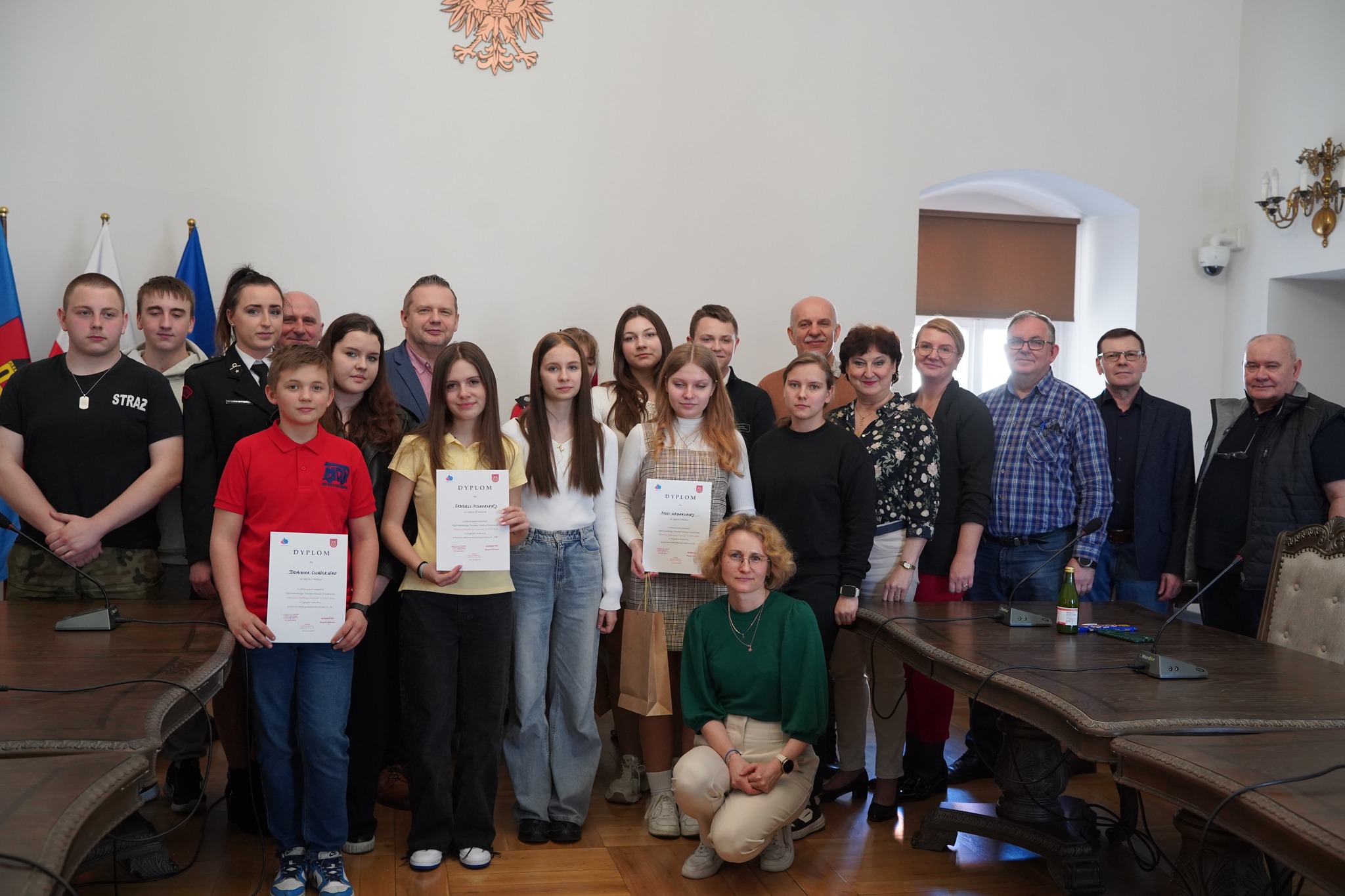 Na zdjęciu widać grupę osób zgromadzoną w sali ratusza miejskiego w Kole. Wśród nich są młodzi uczestnicy trzymający dyplomy, a także organizatorzy i opiekunowie. W tle widoczne są flagi oraz godło Polski. 