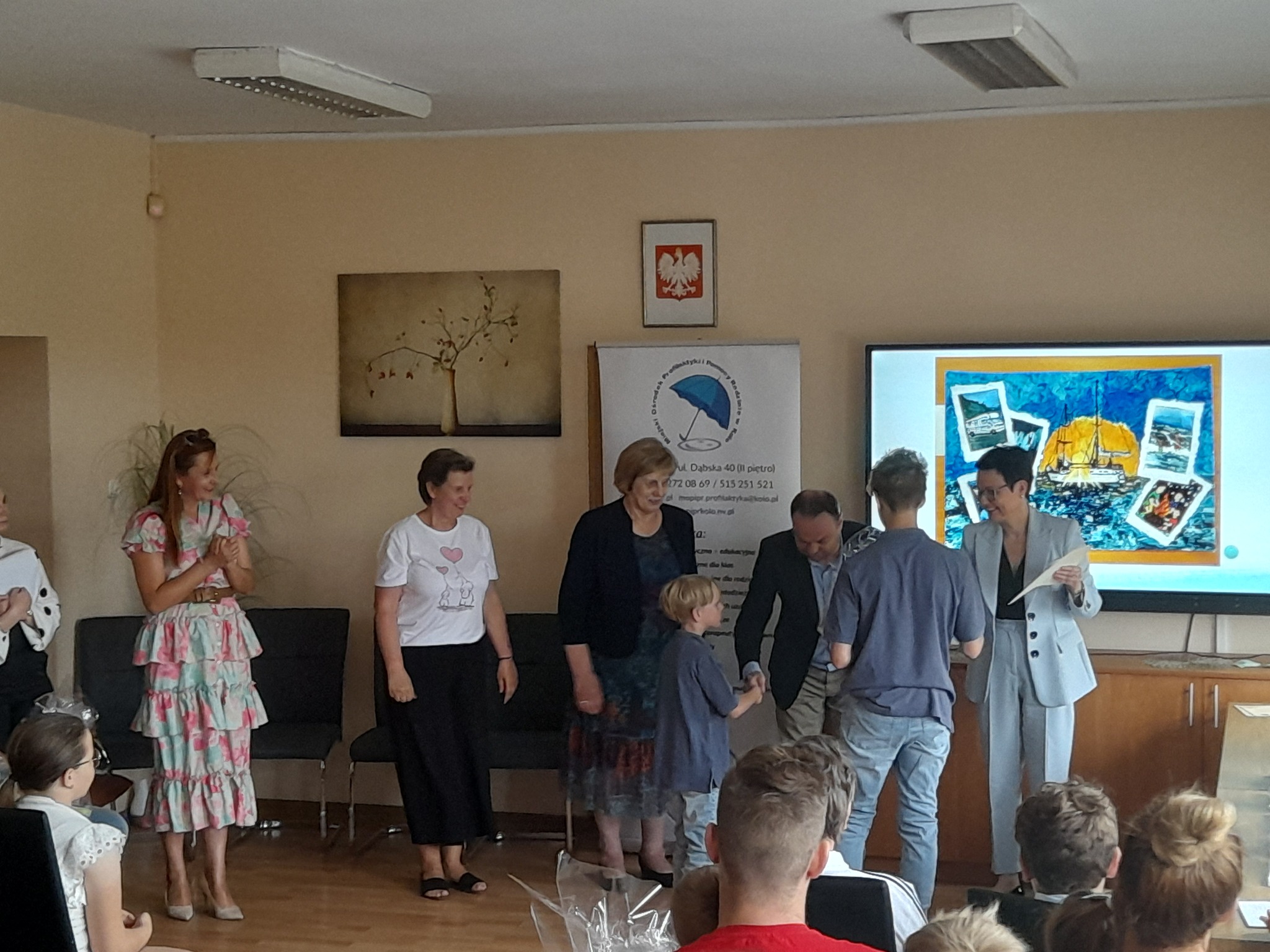 Na zdjęciu stojące uśmiechnięte kobiety, między nimi mężczyzna wręczający dyplom chłopcu