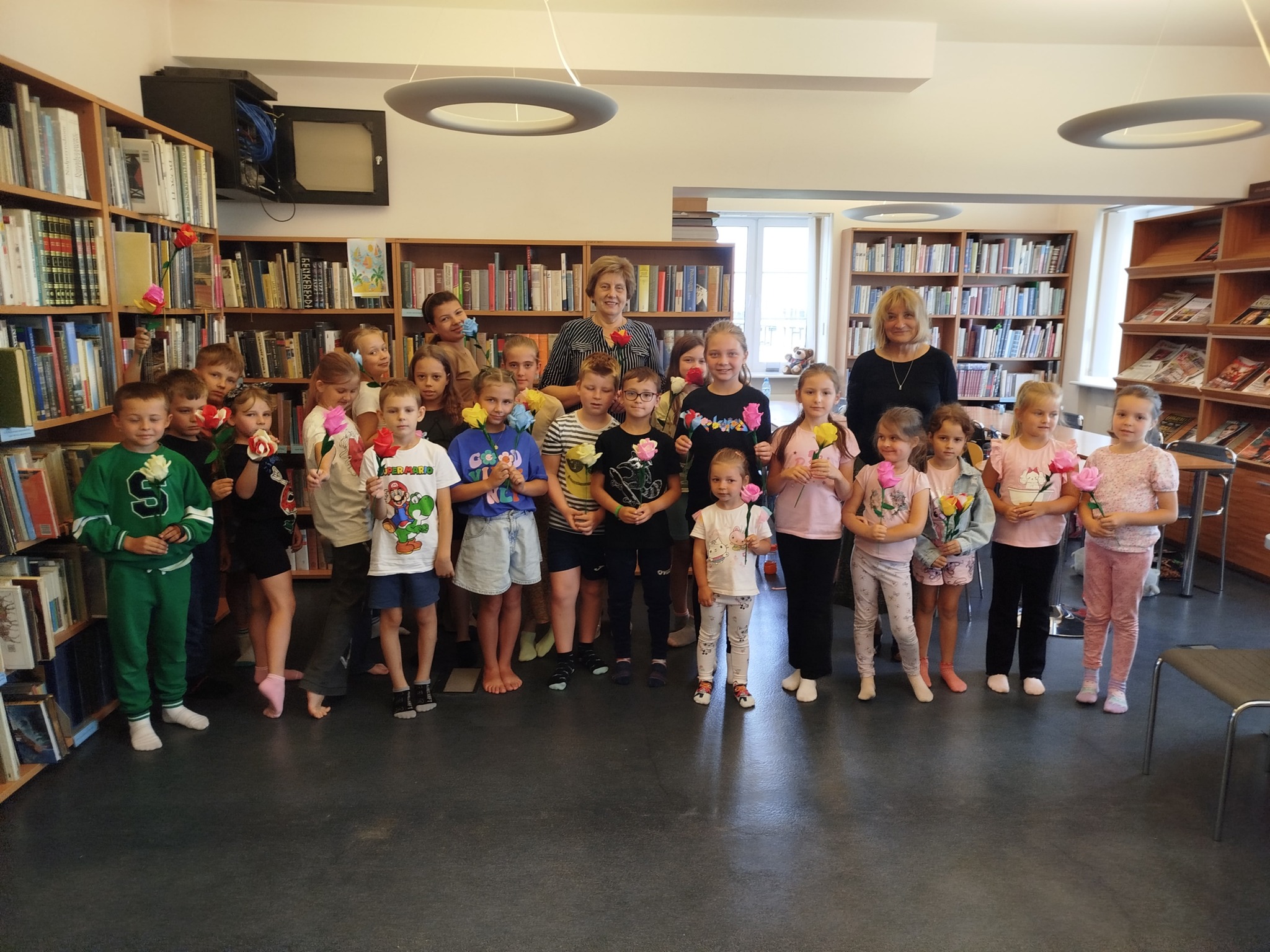 Na zdjęciu widać grupę dzieci i dwie dorosłe kobiety w bibliotece, wszyscy trzymają kolorowe kwiaty z papieru – prawdopodobnie po zakończonych warsztatach plastycznych.